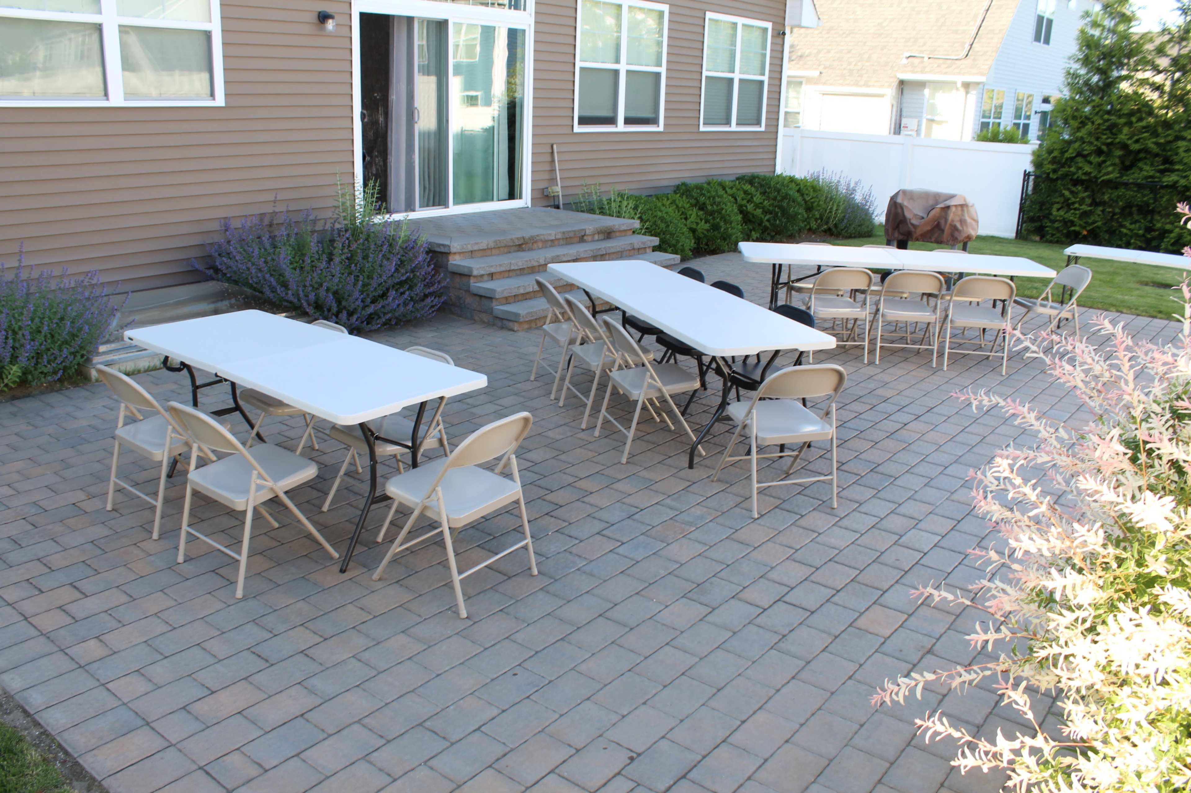 A group of folding tables and chairs is arranged on a paved patio outside a house.