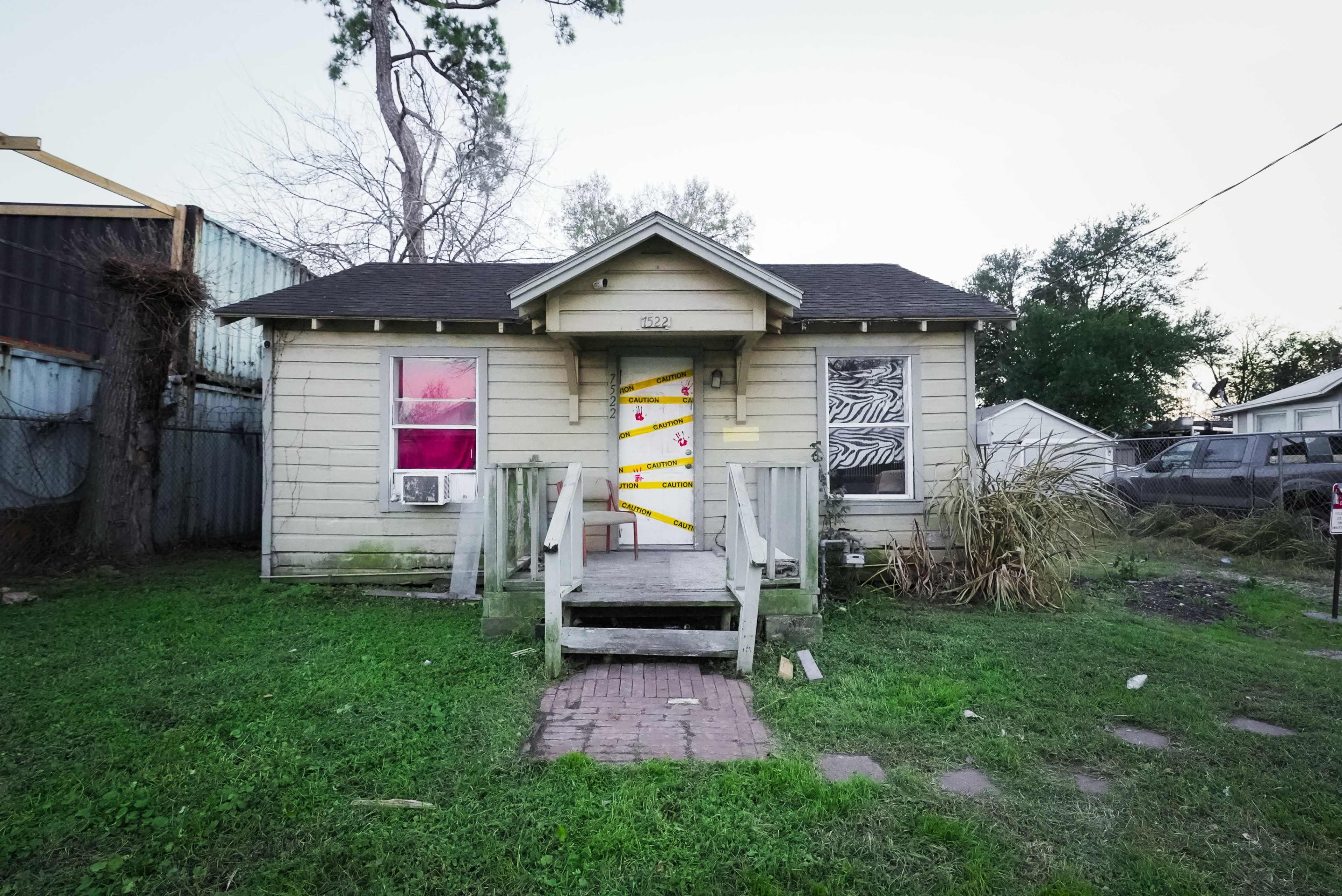 The image shows a small wooden house with a ramp leading to a front door, featuring colorful decorations and a partially overgrown yard.
