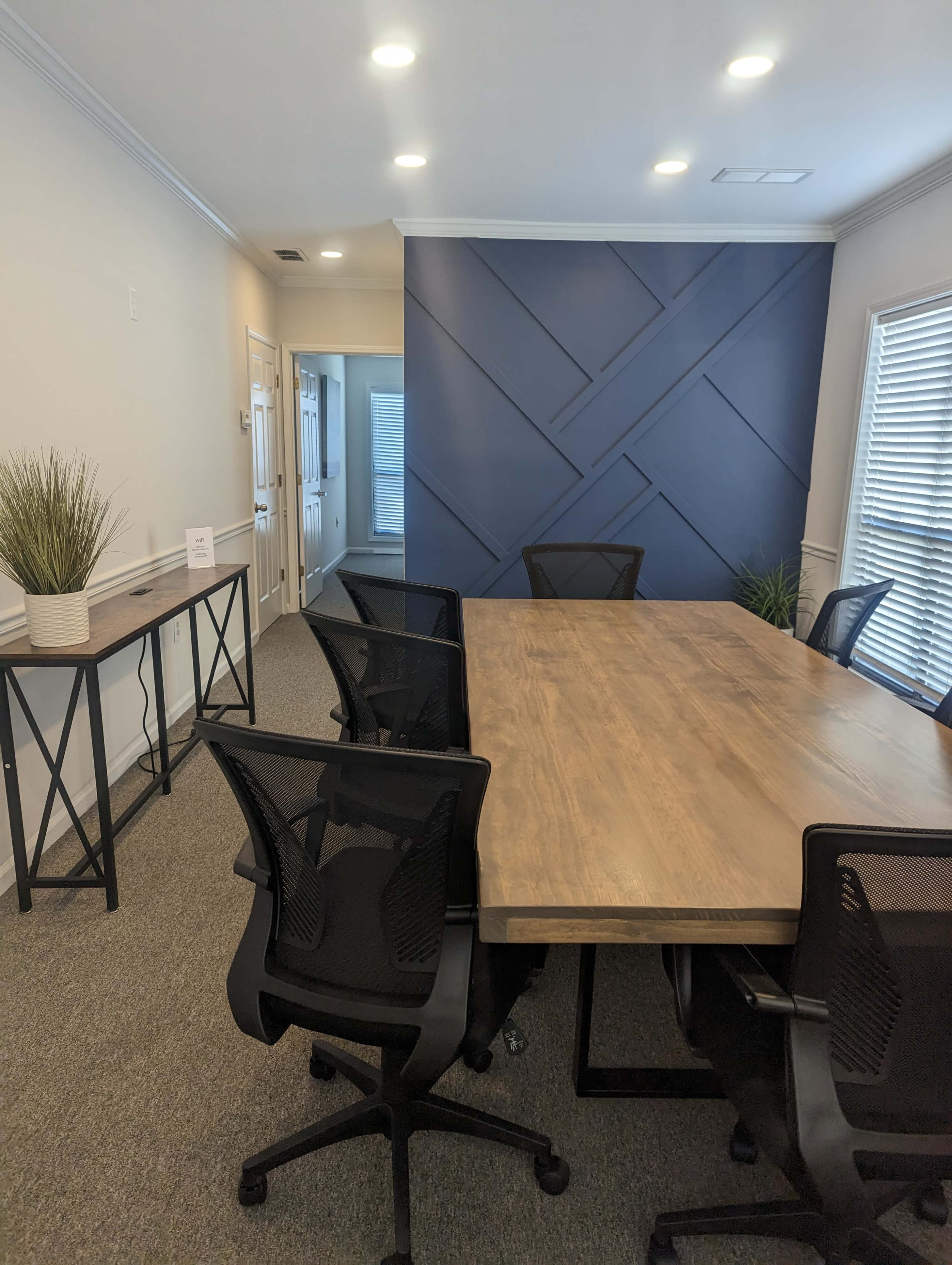 A conference room with a wooden table surrounded by black mesh chairs, featuring a blue accent wall and potted plants.