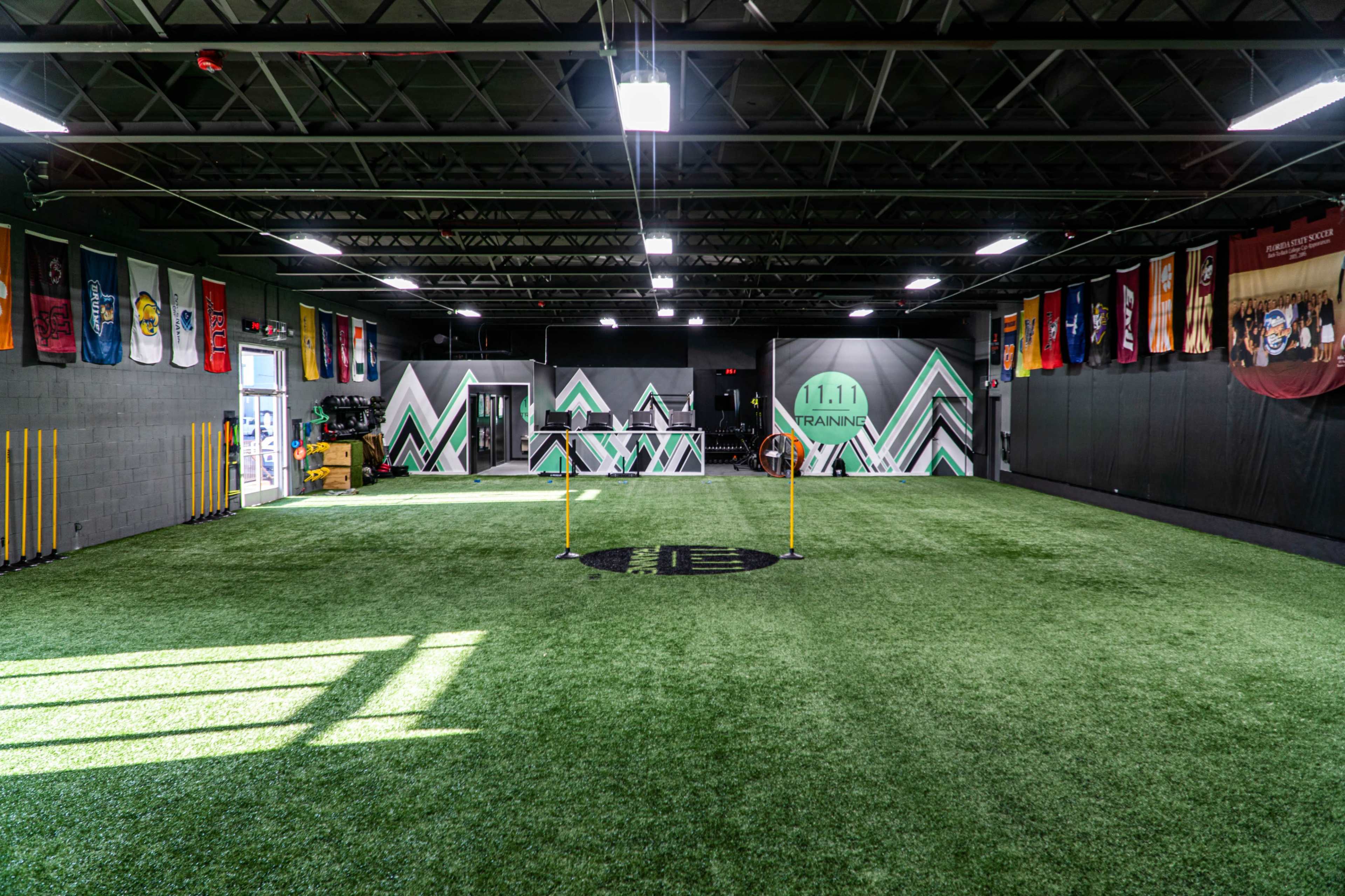 An indoor training facility with artificial turf, mirrored walls, and various sports flags displayed.