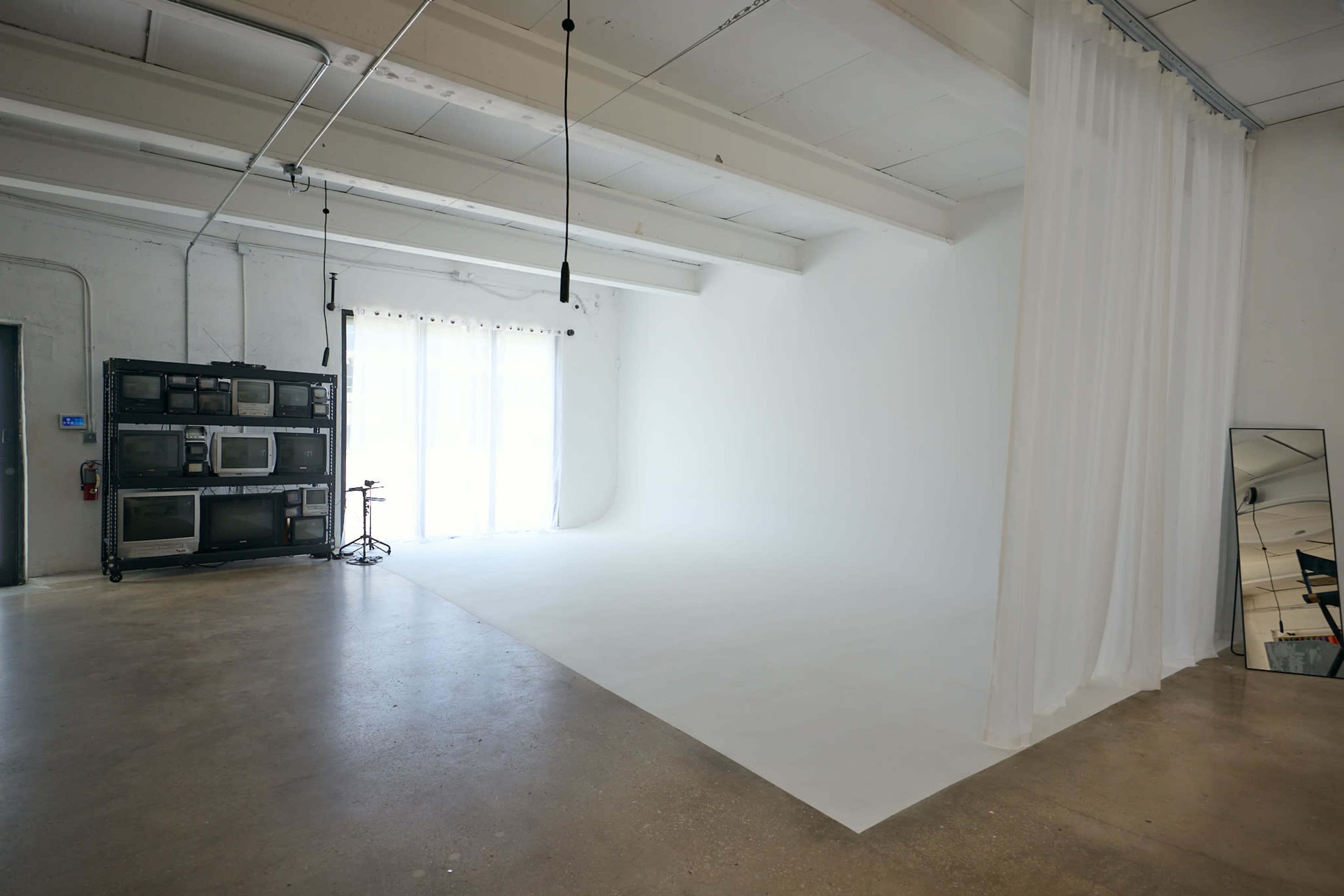 The image shows an empty studio space with a large white backdrop, natural light coming through window curtains, and minimal furnishings along one wall.