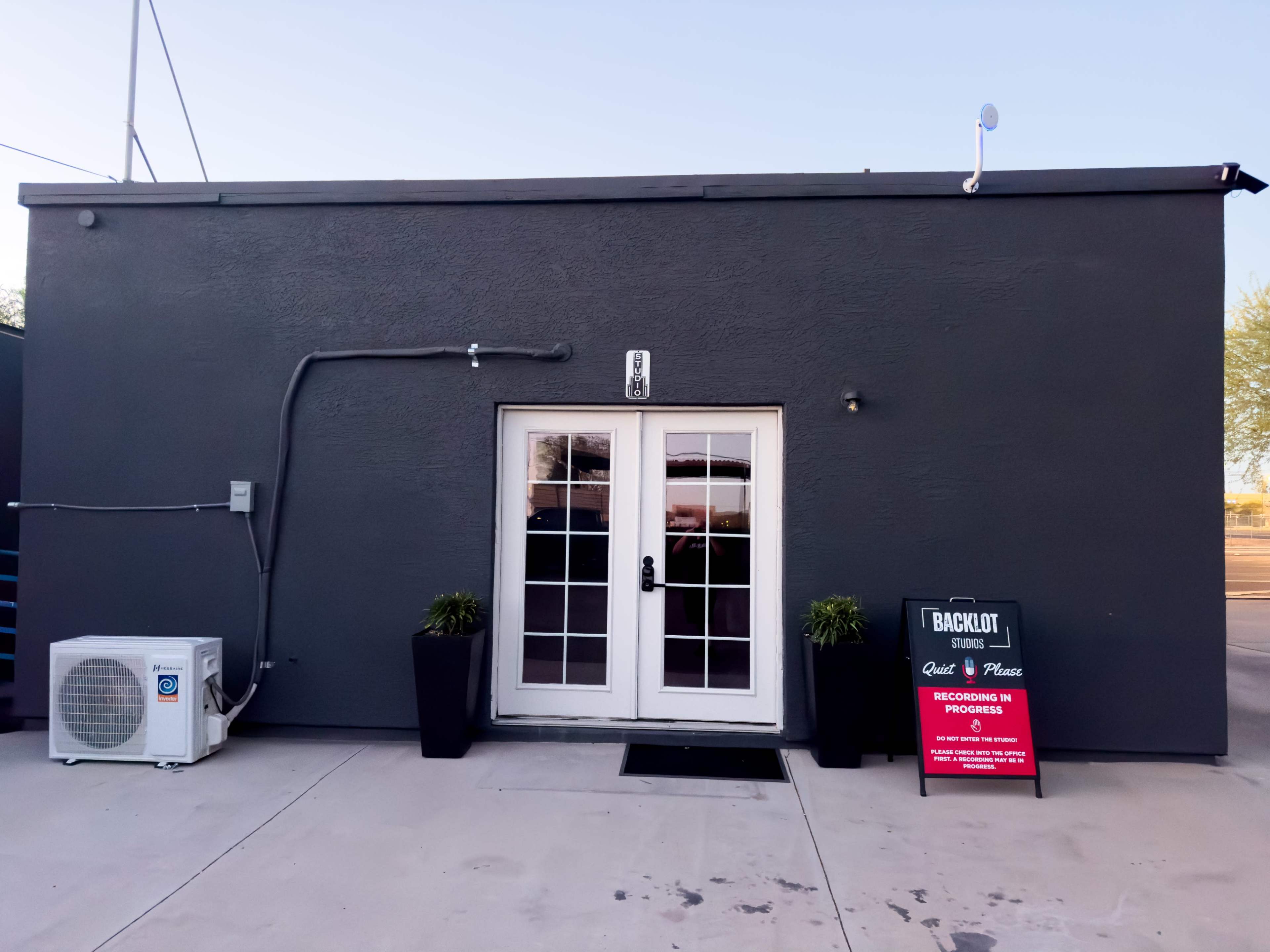 The image shows a gray building with a double-door entrance and a sign on a nearby stand.