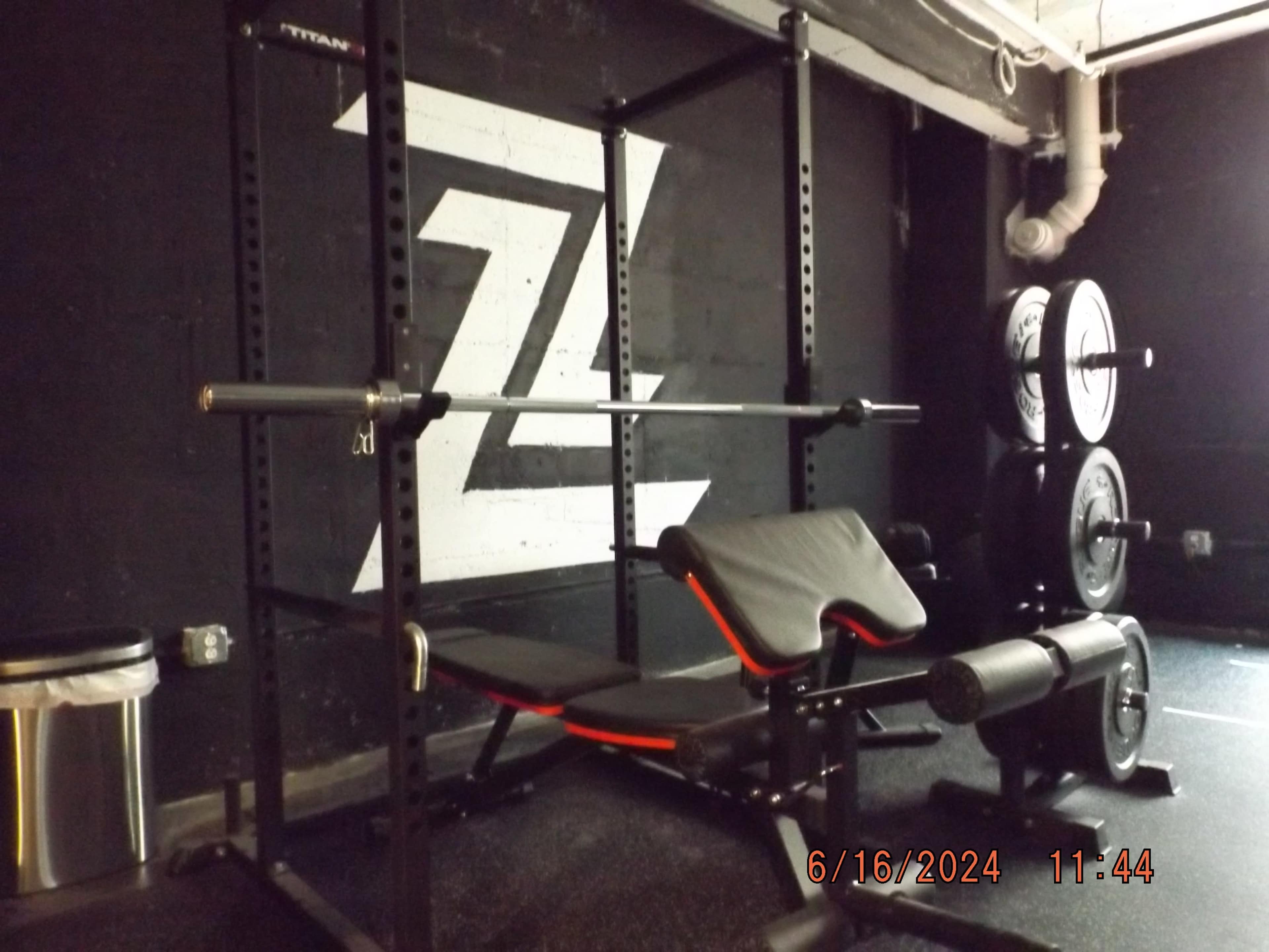 The image shows a gym setup featuring a squat rack, a bench with an adjustable seat, and a set of weight plates stacked on a rack against a black wall.