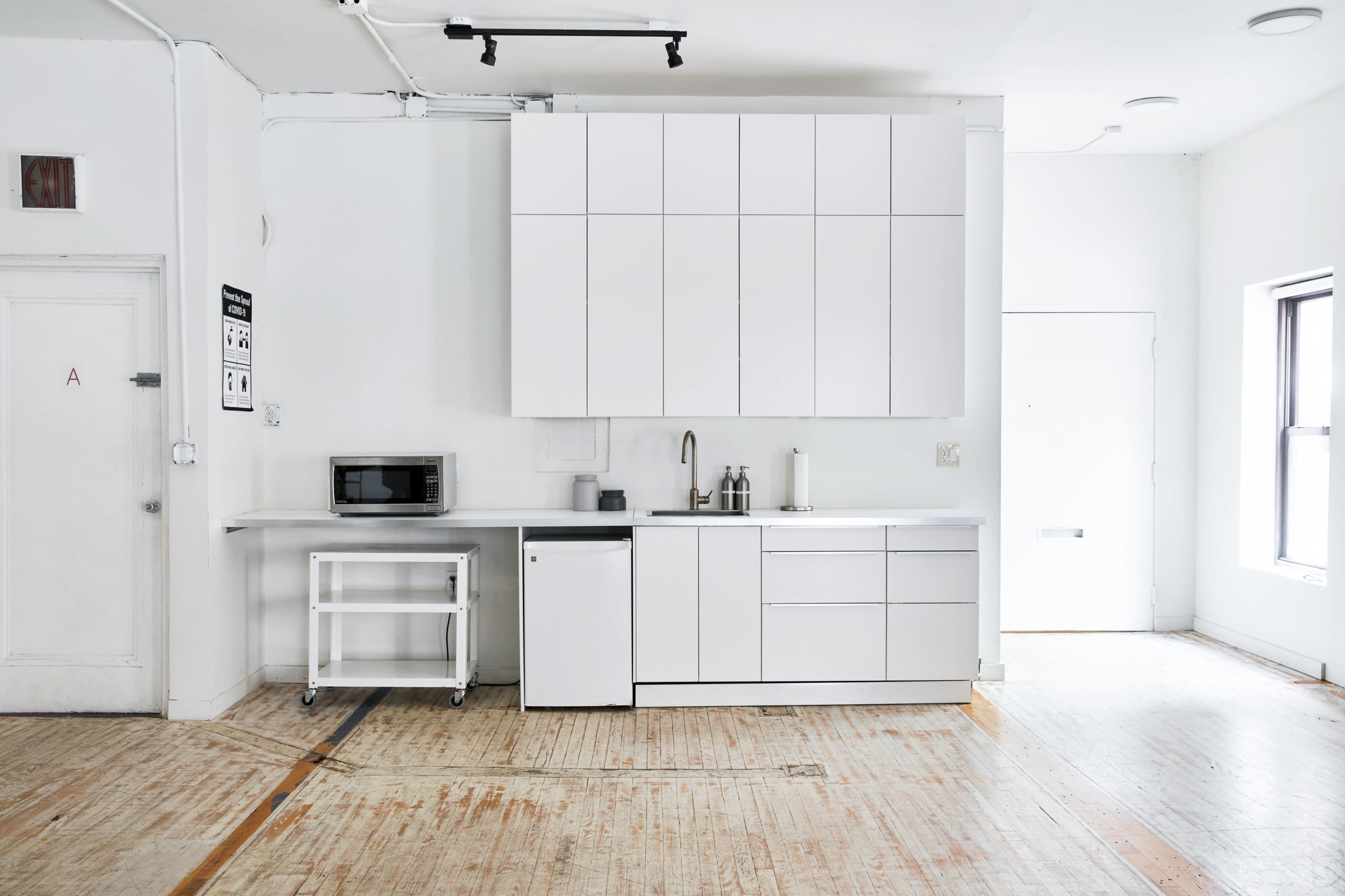 A minimalist kitchen with white cabinetry, a microwave, a small refrigerator, and an empty space on a wooden floor.