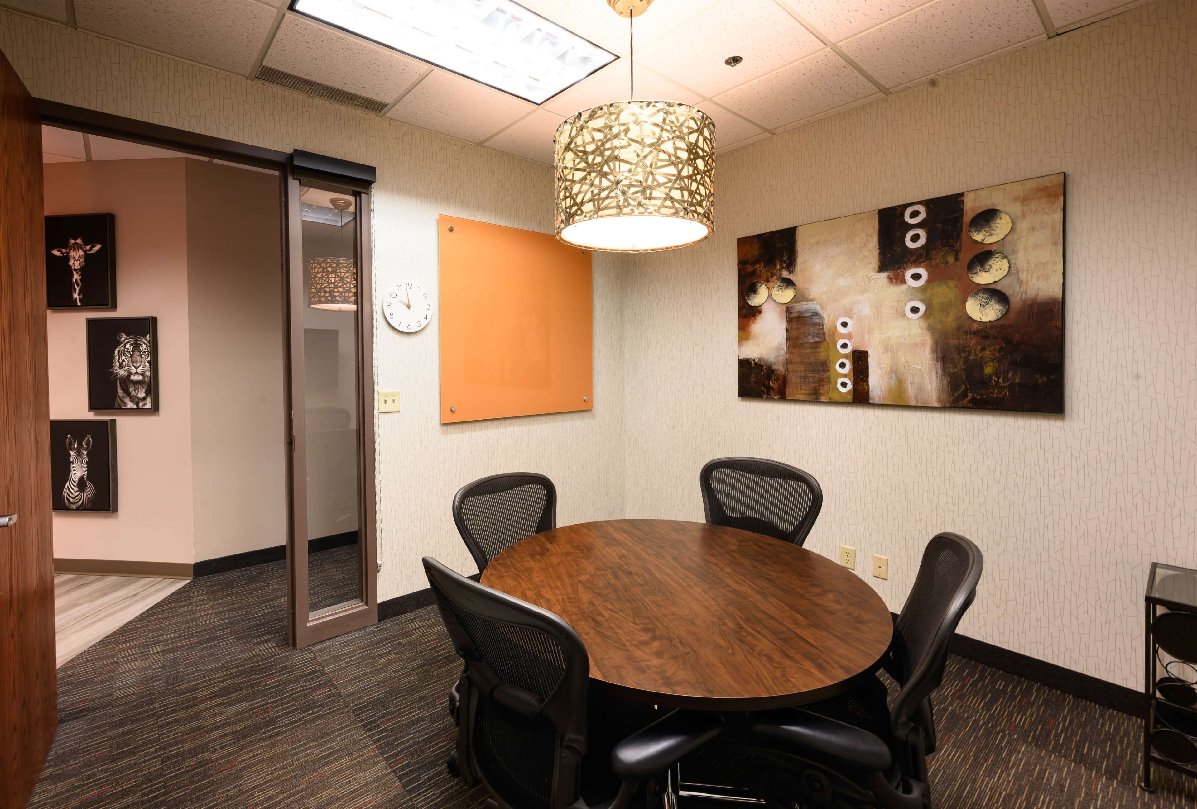 The image shows a small conference room with a circular wooden table surrounded by black mesh chairs, featuring an abstract wall painting and a decorative light fixture.