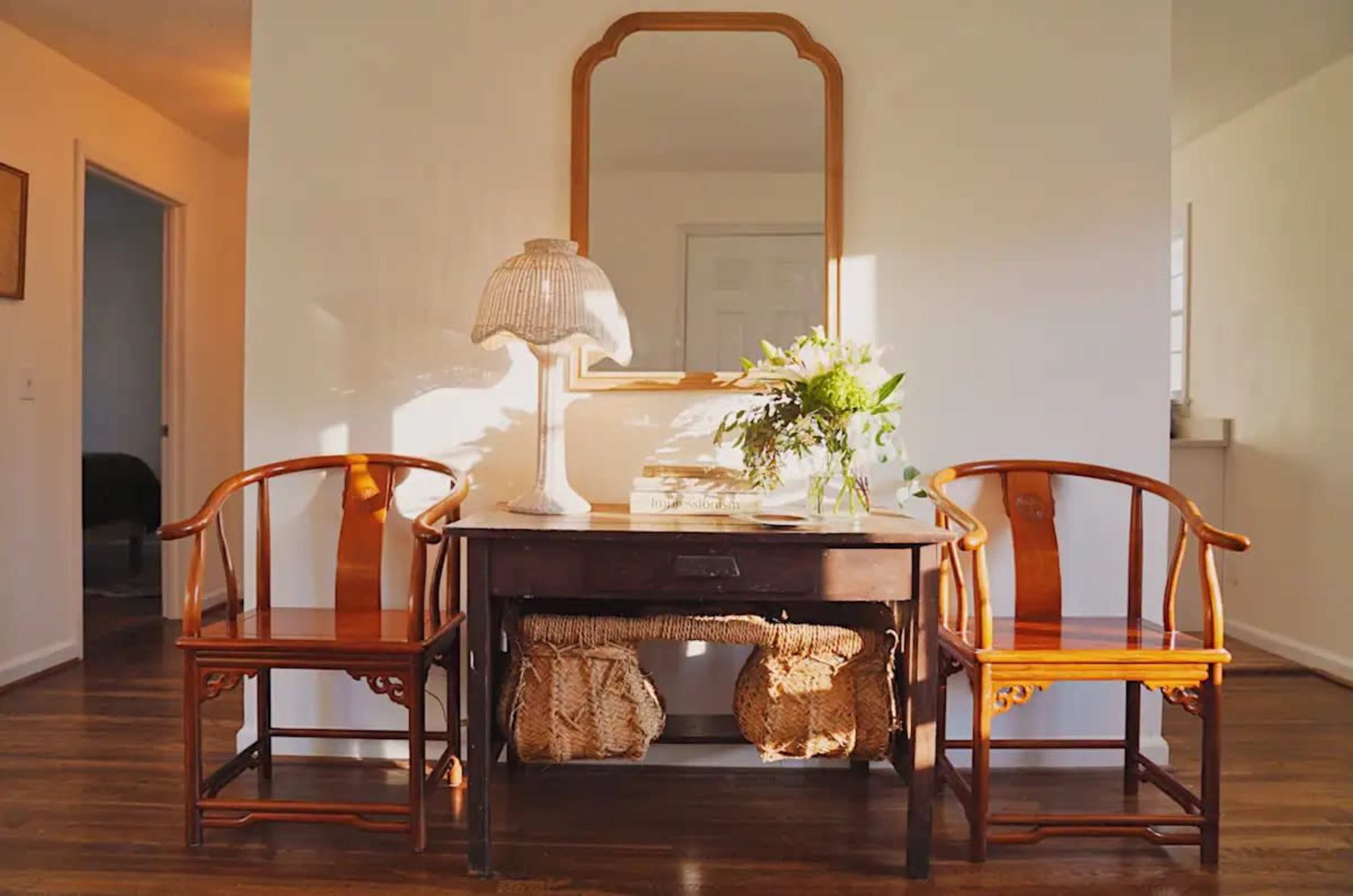 A wooden table with a mirror above it is flanked by two chairs and a lamp, with a vase of flowers placed on the table.