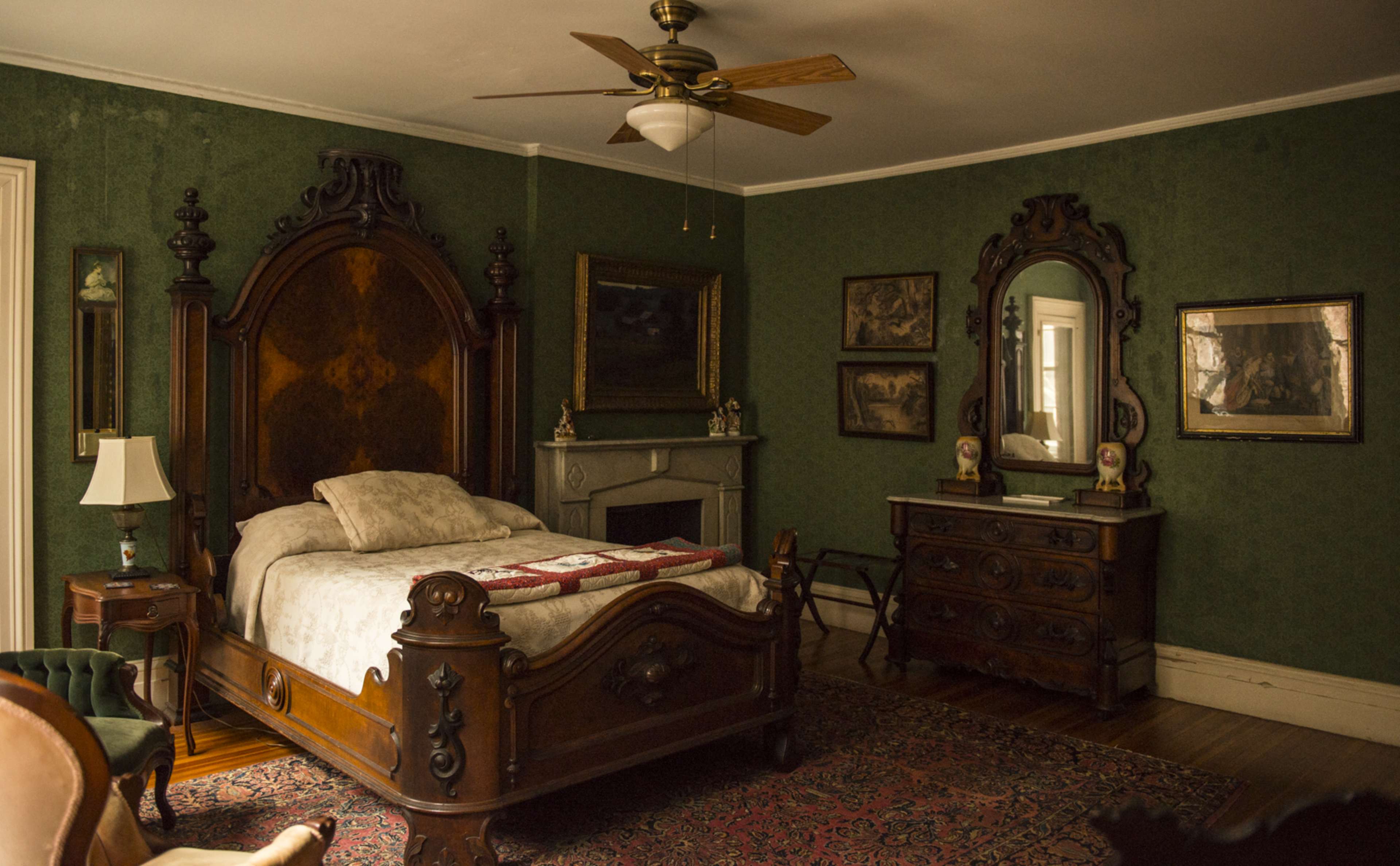 The room features a large wooden bed with an ornate headboard, a fireplace with a mantel, a dresser with a mirror, and green wallpaper adorned with paintings.