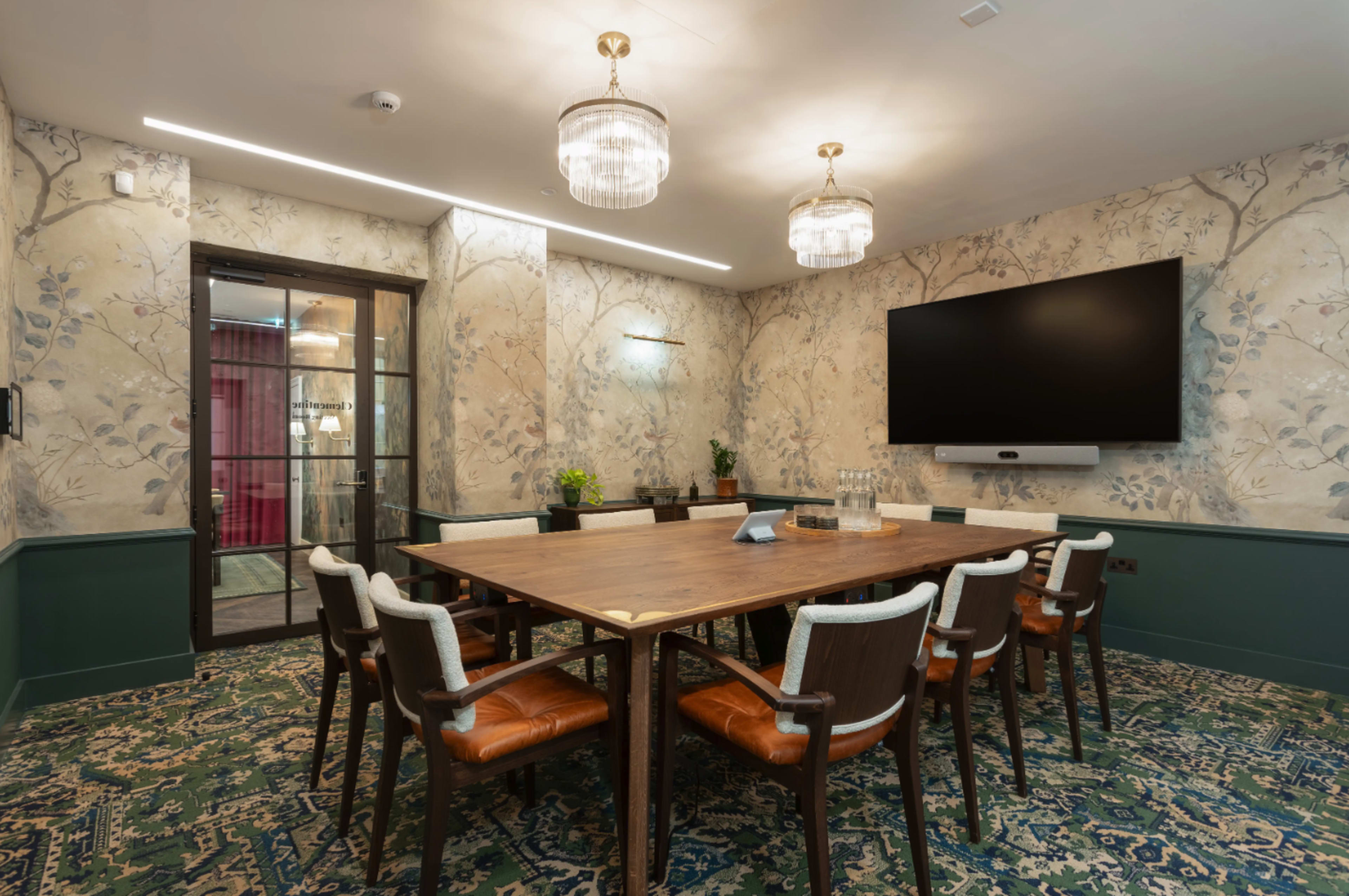 The image shows a conference room featuring a large wooden table surrounded by leather and upholstered chairs, with floral wallpaper and a large screen mounted on the wall.