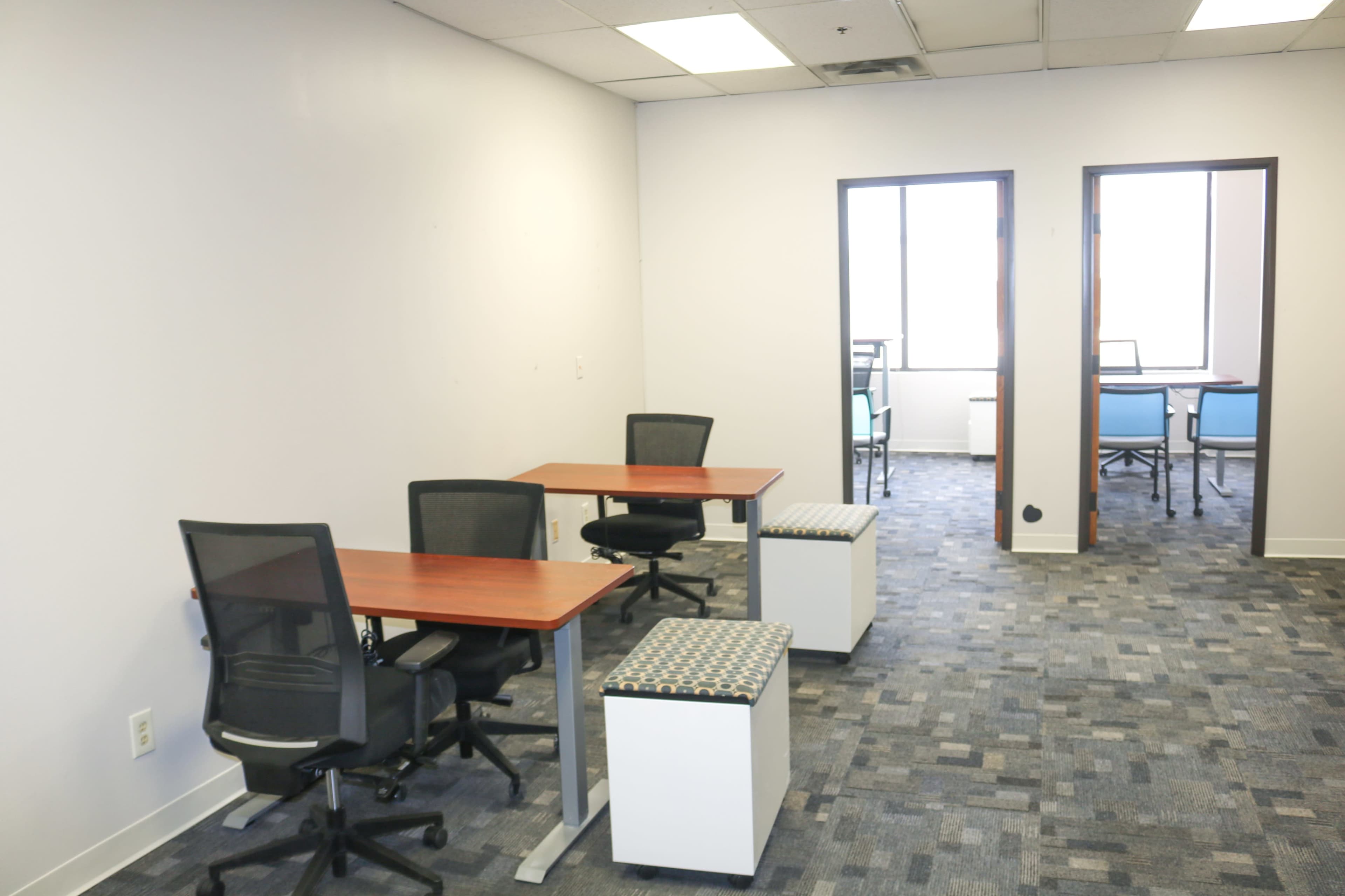 The image shows a modern office space with empty desks, chairs, and a carpeted floor.