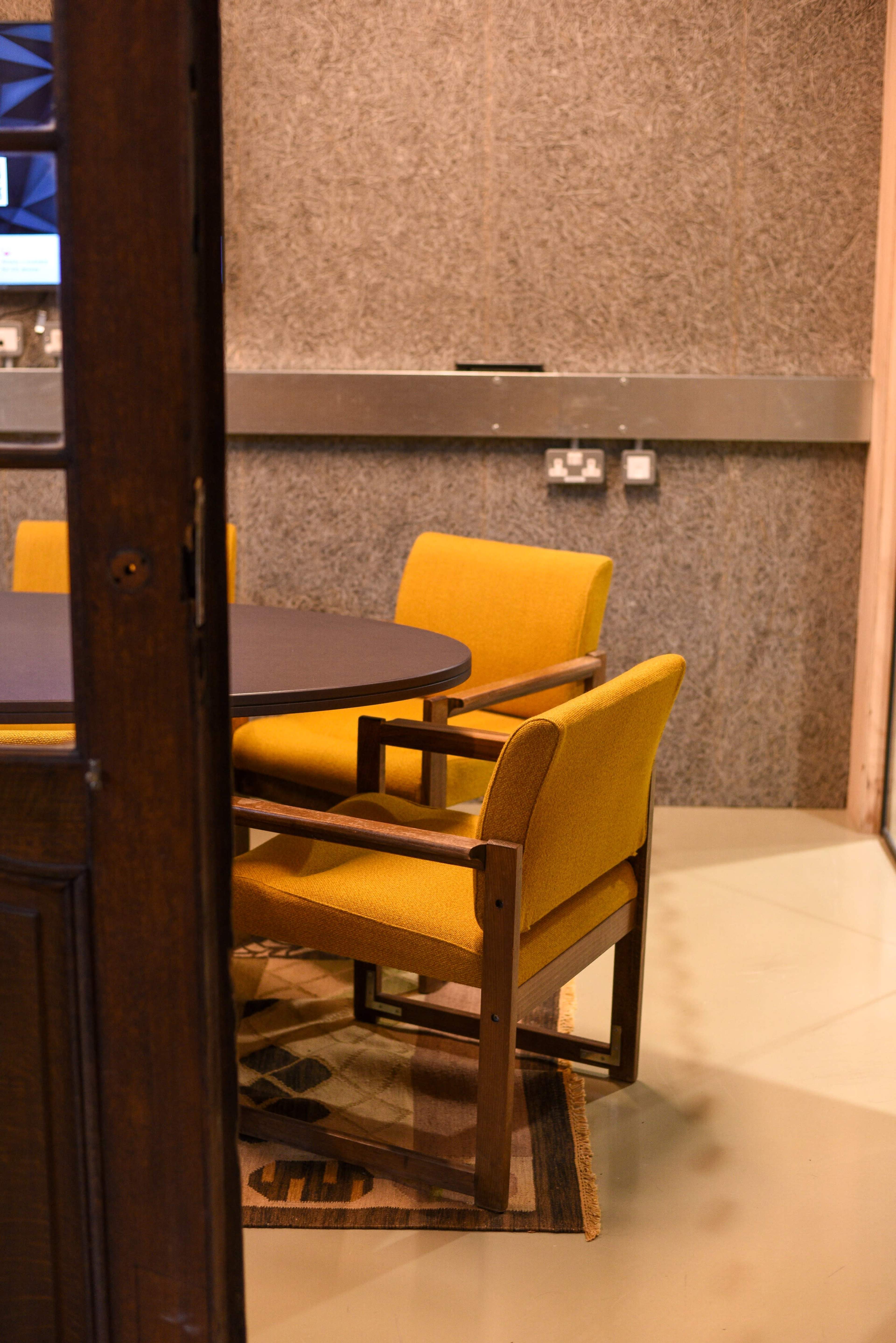 A wooden door partially opens to reveal a round table with yellow upholstered chairs on a patterned rug in a room with textured gray walls.