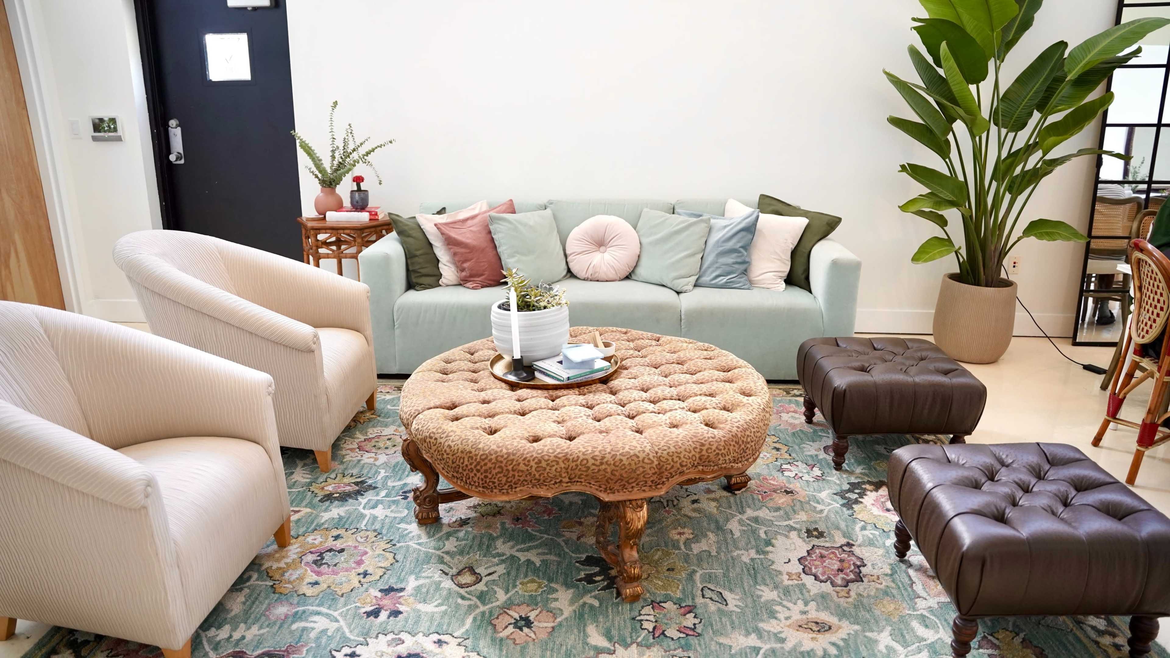 A light-colored living room features a tufted ottoman at the center, surrounded by a pastel couch, two armchairs, and a potted plant.