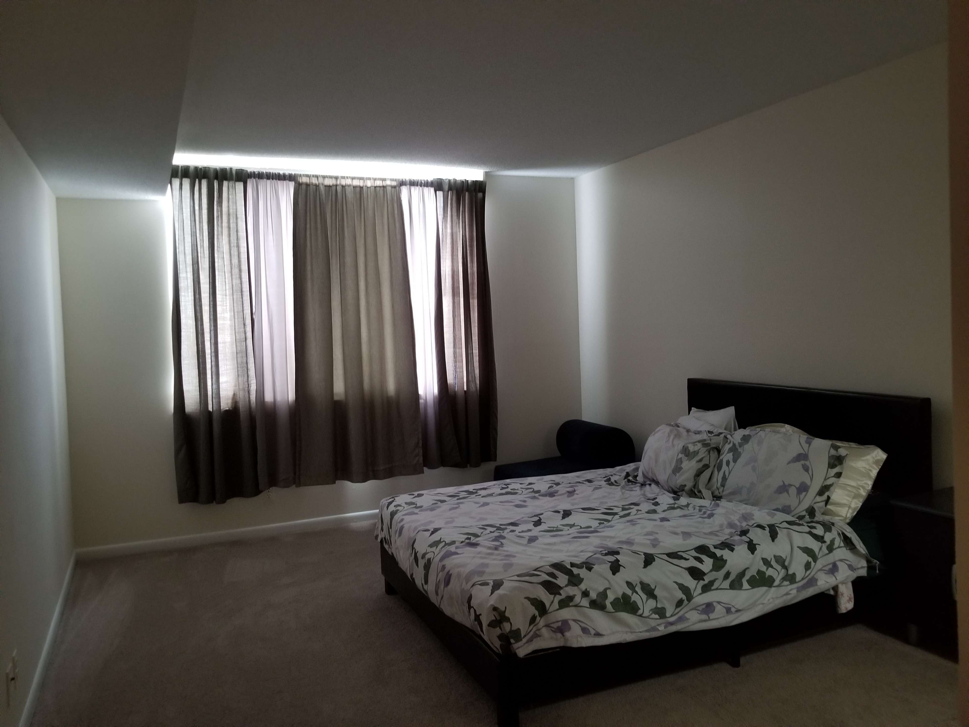 A neatly made bed with a floral-patterned blanket is positioned against a wall under a large window with dark curtains in a sparsely furnished bedroom.