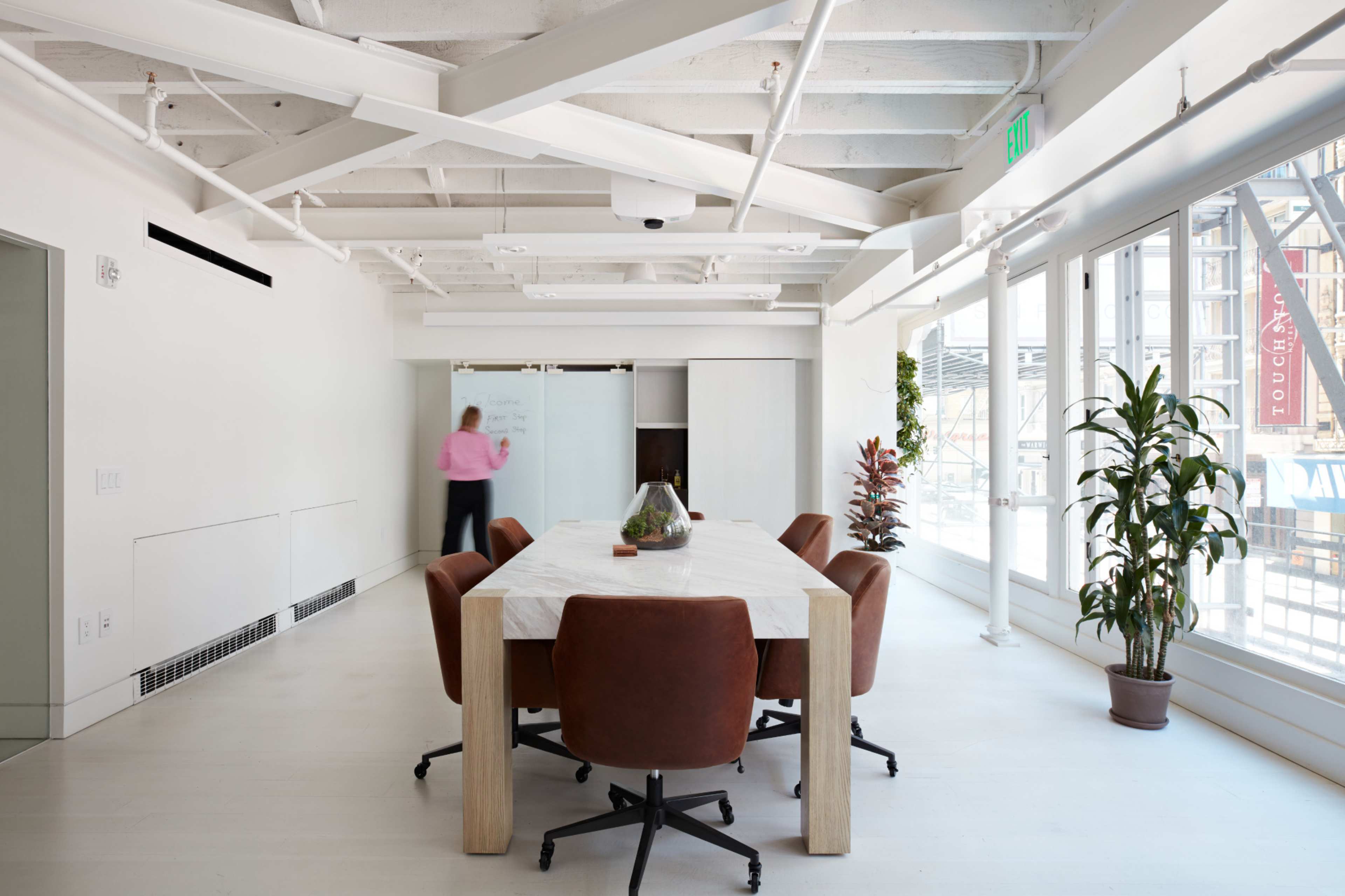 A modern conference room features a large white table surrounded by brown chairs, with a glass wall allowing natural light and visibility to an outside area.