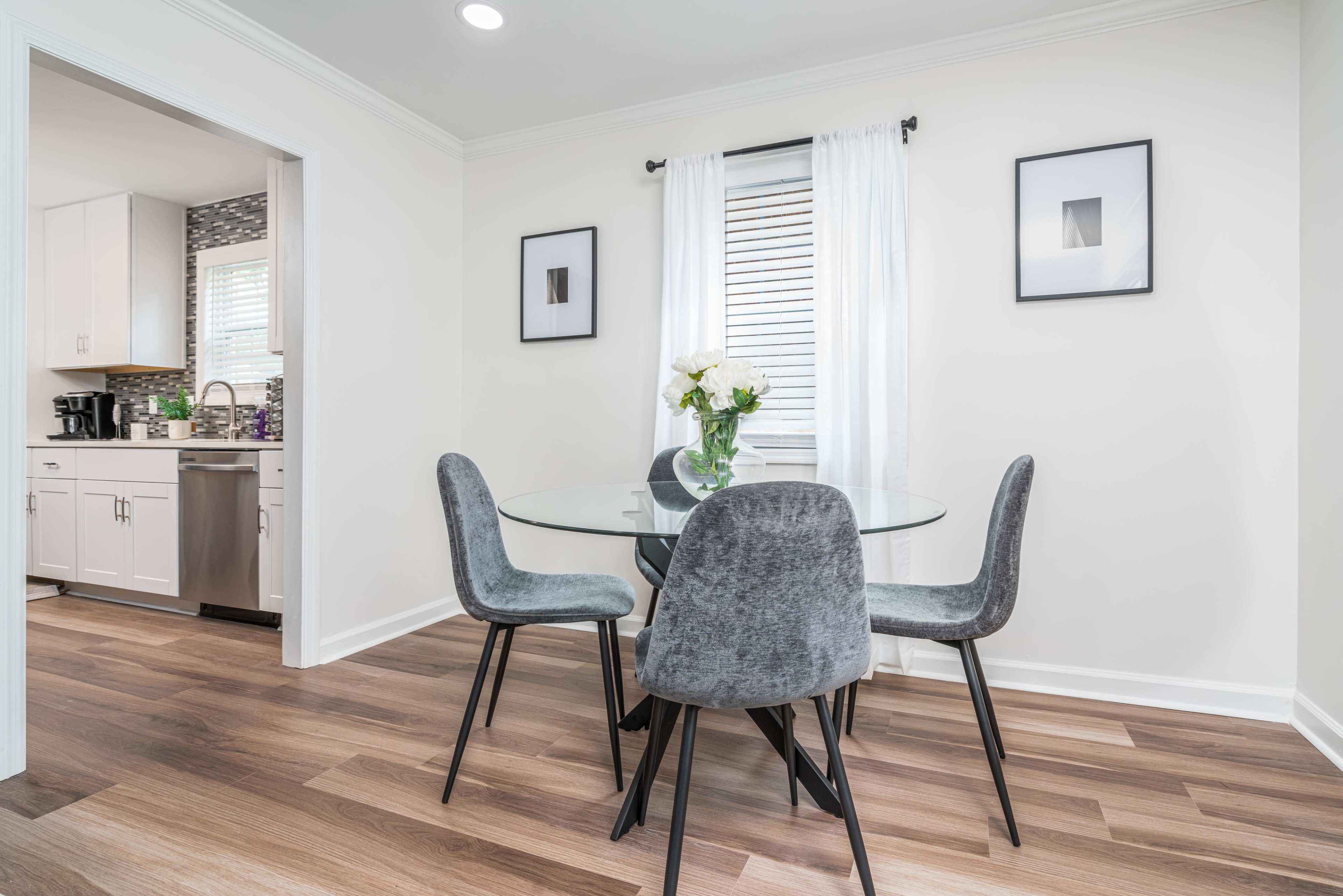 A round glass dining table with four gray chairs is set in a bright, minimalist kitchen space featuring white walls and wood flooring.
