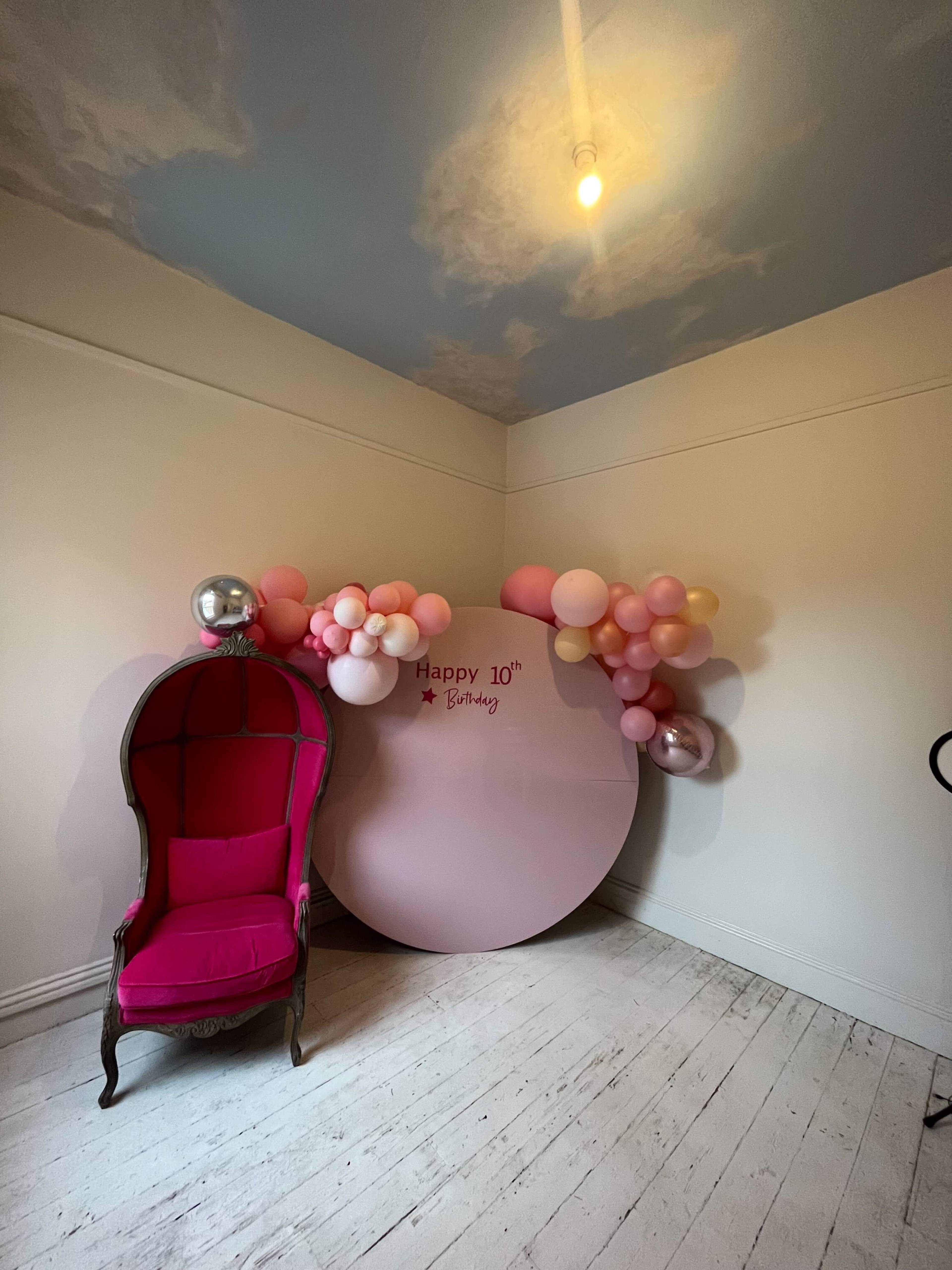 A pink chair is positioned next to a large circular backdrop that reads "Happy 10th Birthday," adorned with a cluster of pink and white balloons.
