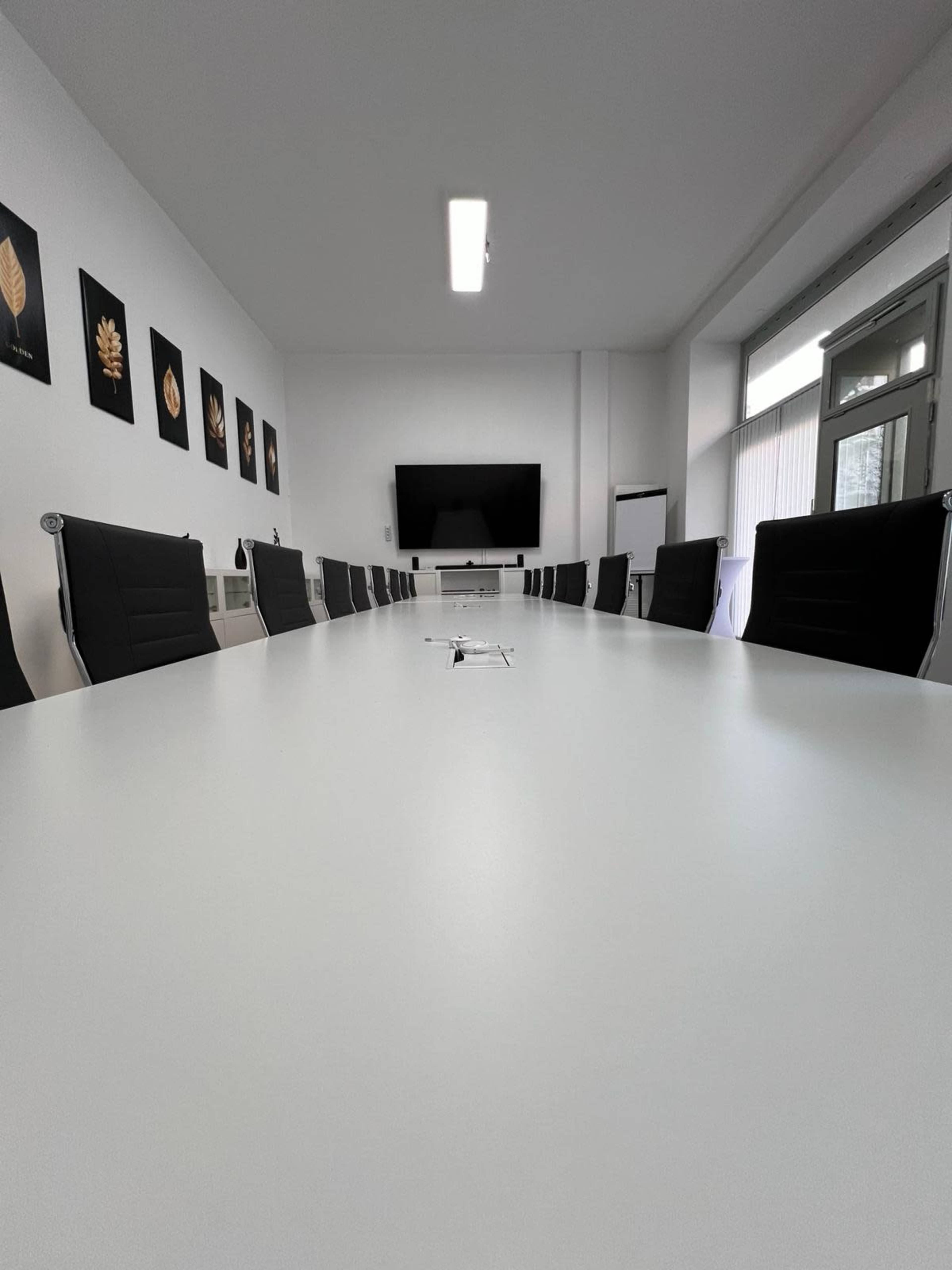 A large, white conference table is set in a minimalist meeting room with black chairs and a wall-mounted screen at one end.