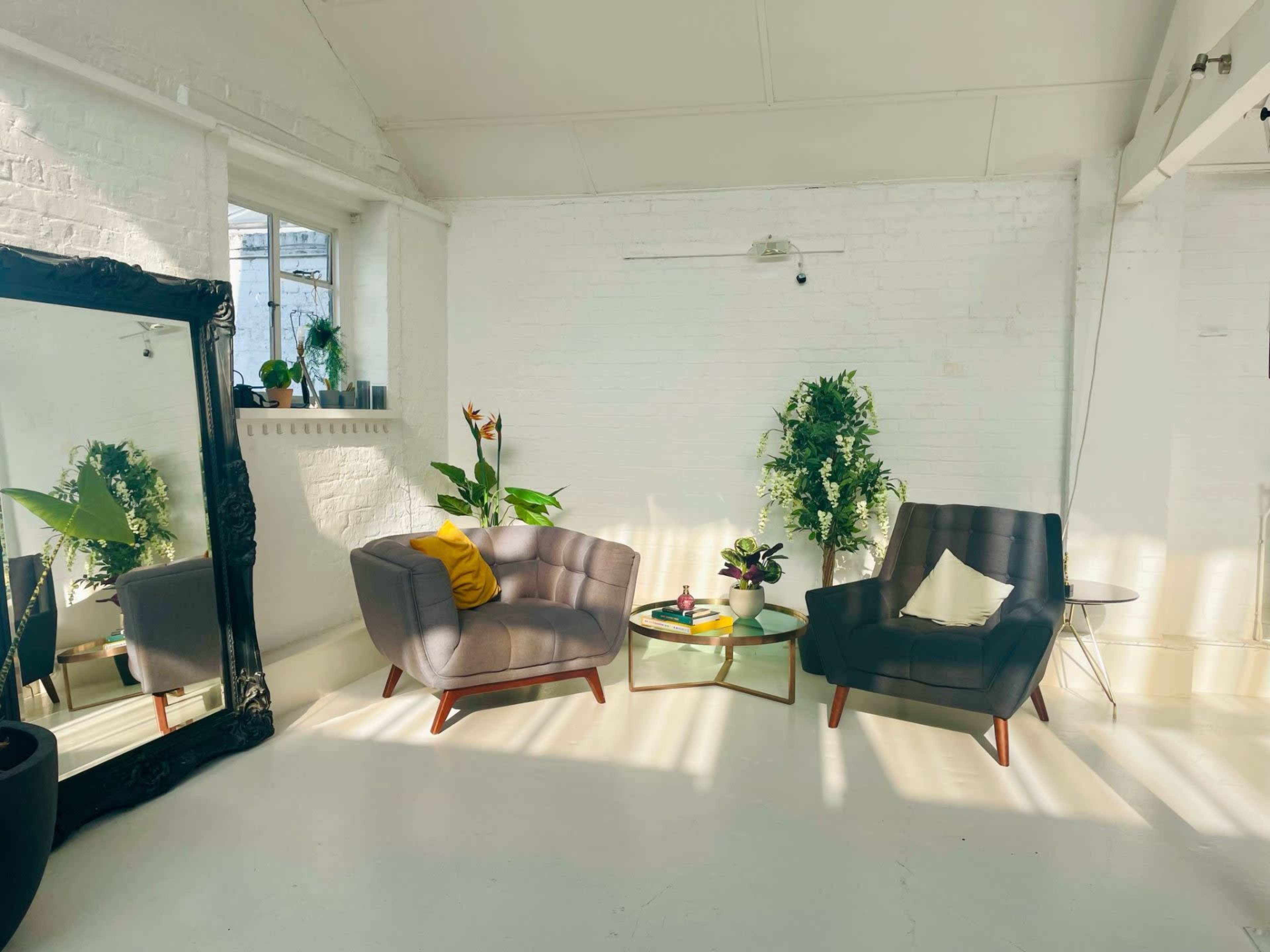 The image shows a bright, minimalist interior featuring two armchairs, a small glass table with plants, and a large mirror reflecting the space.