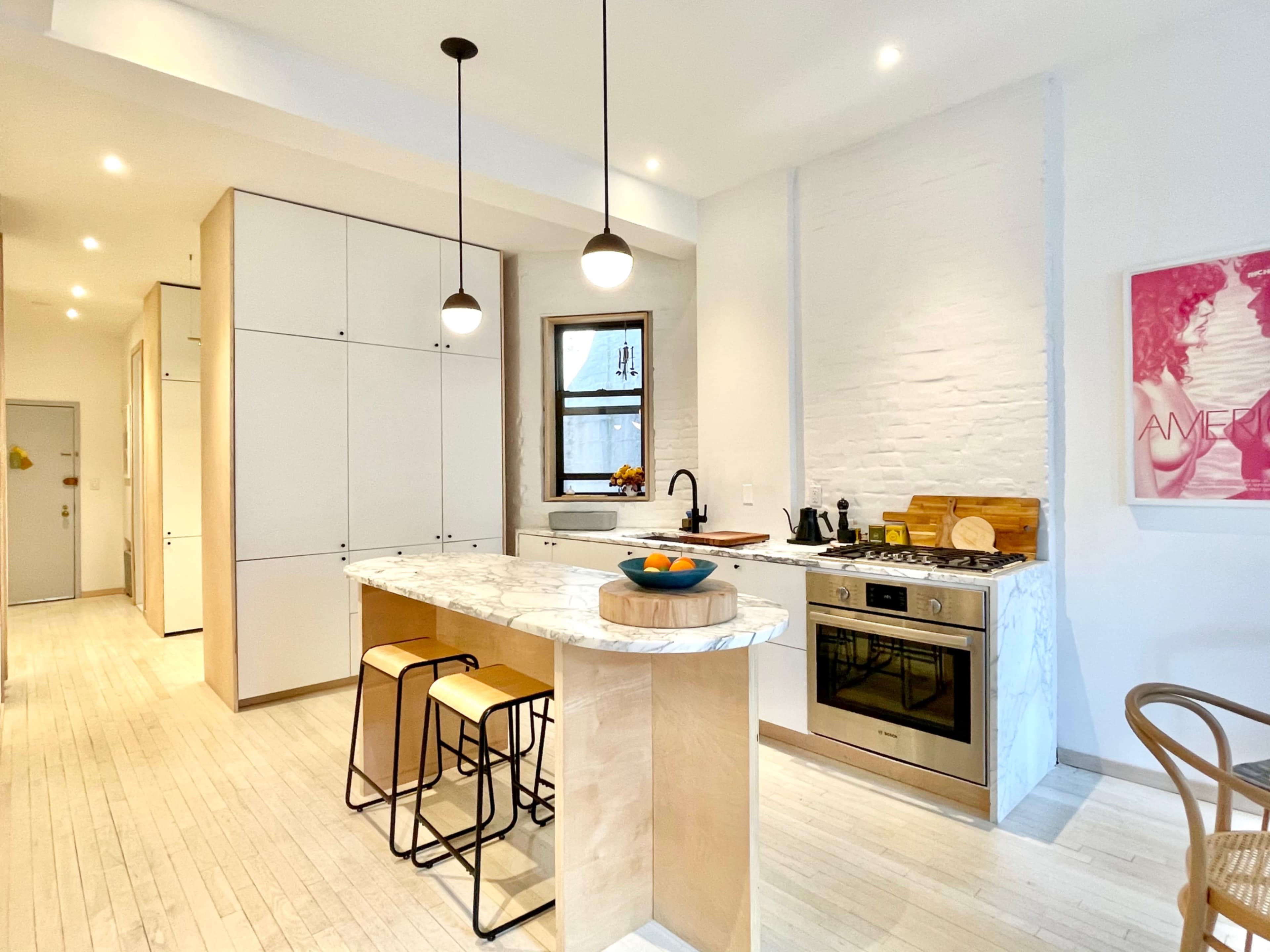 A modern kitchen features a marble island with bar stools, stainless steel appliances, and white cabinetry, illuminated by pendant lights.