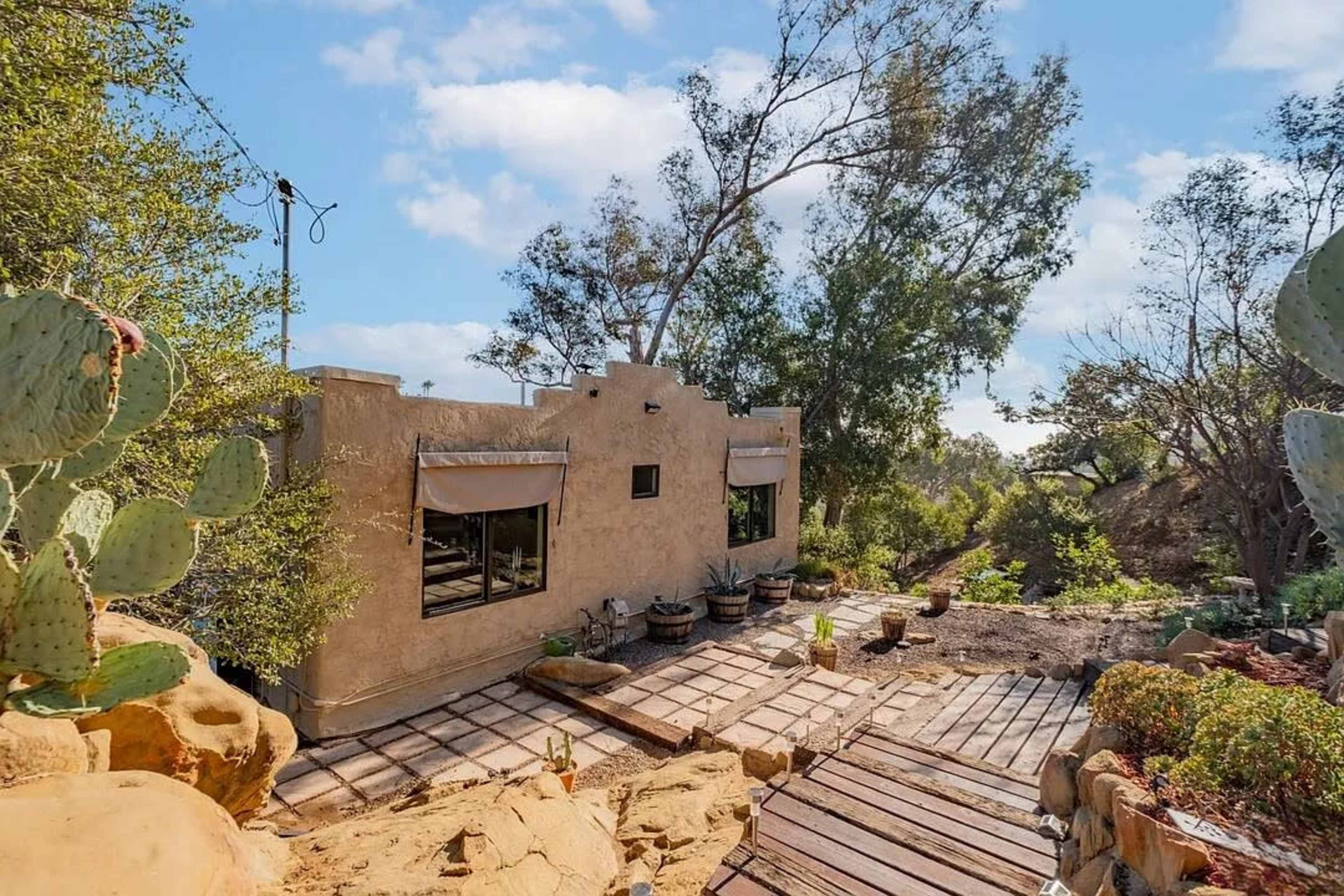 A rustic, earthen-colored building with large windows and a wooden deck is surrounded by desert vegetation and trees under a clear blue sky.