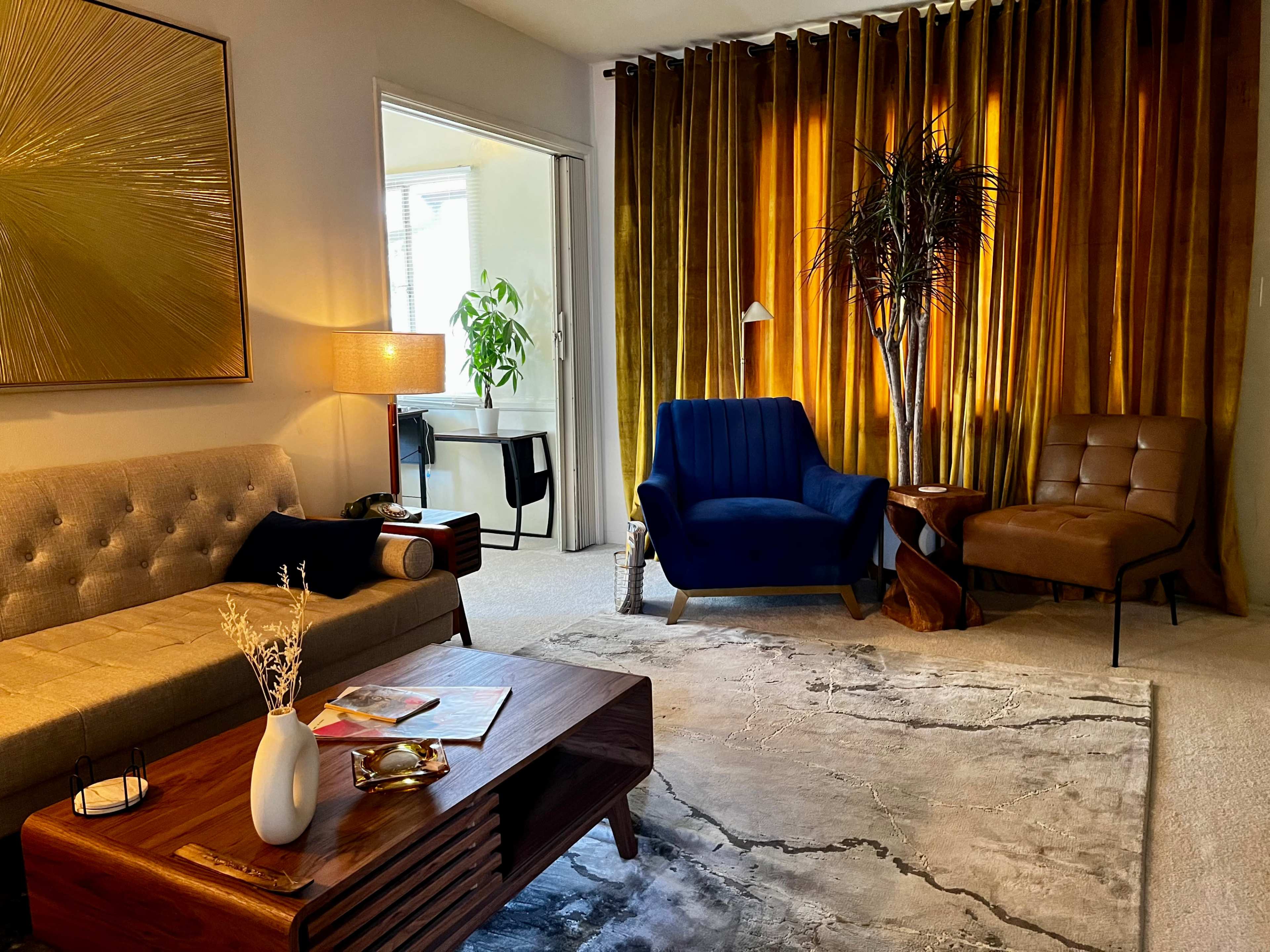 A well-furnished living room with a gray sofa, a blue armchair, a brown chair, a wooden coffee table, and a backdrop of golden curtains.