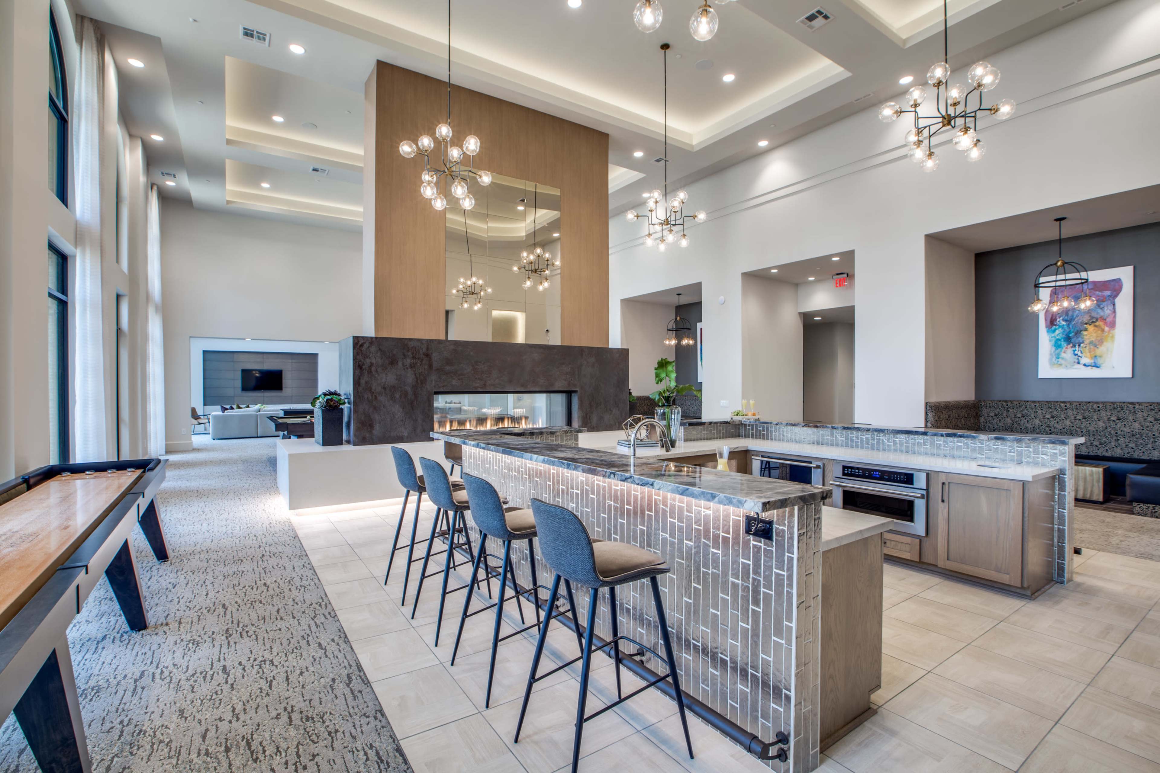 The image shows a modern community lounge area with a kitchenette, a fireplace, and seating arrangements, featuring sleek design elements and ample natural light.