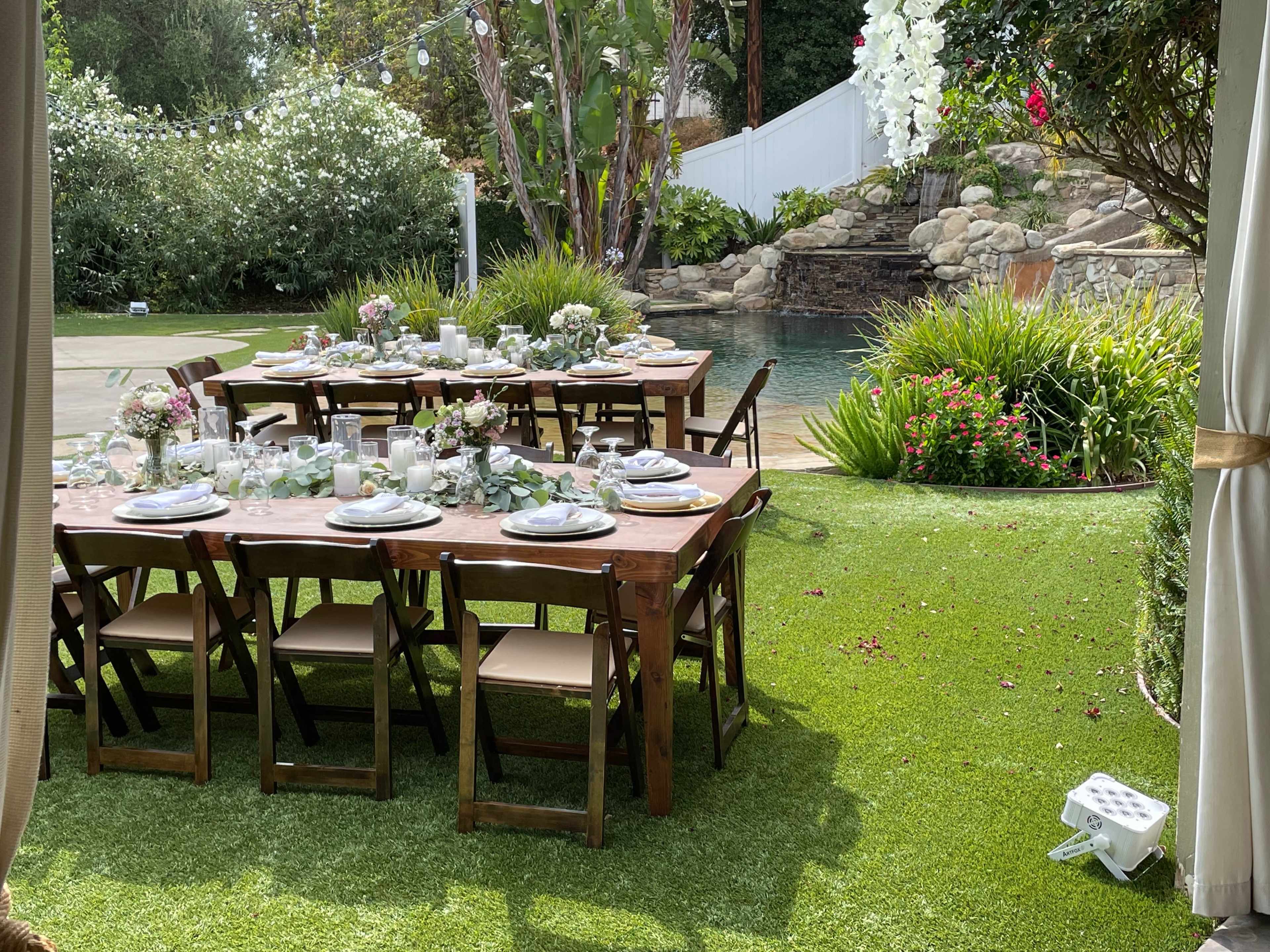 A long wooden dining table is set with plates and centerpieces in a lush garden near a pool.