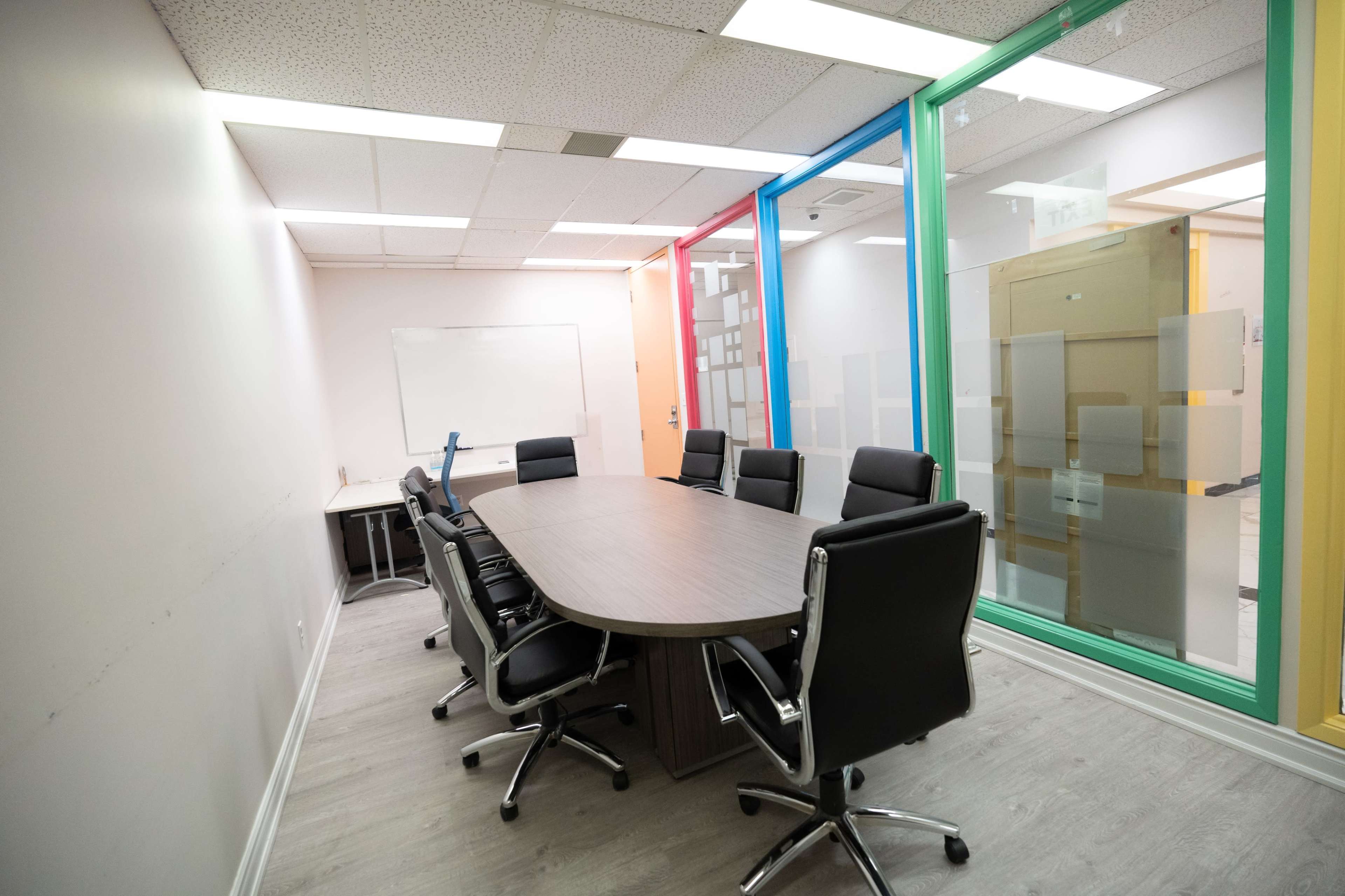 A conference room features a large oval table surrounded by black office chairs, with a whiteboard on one wall and glass panels in vibrant colors on another.