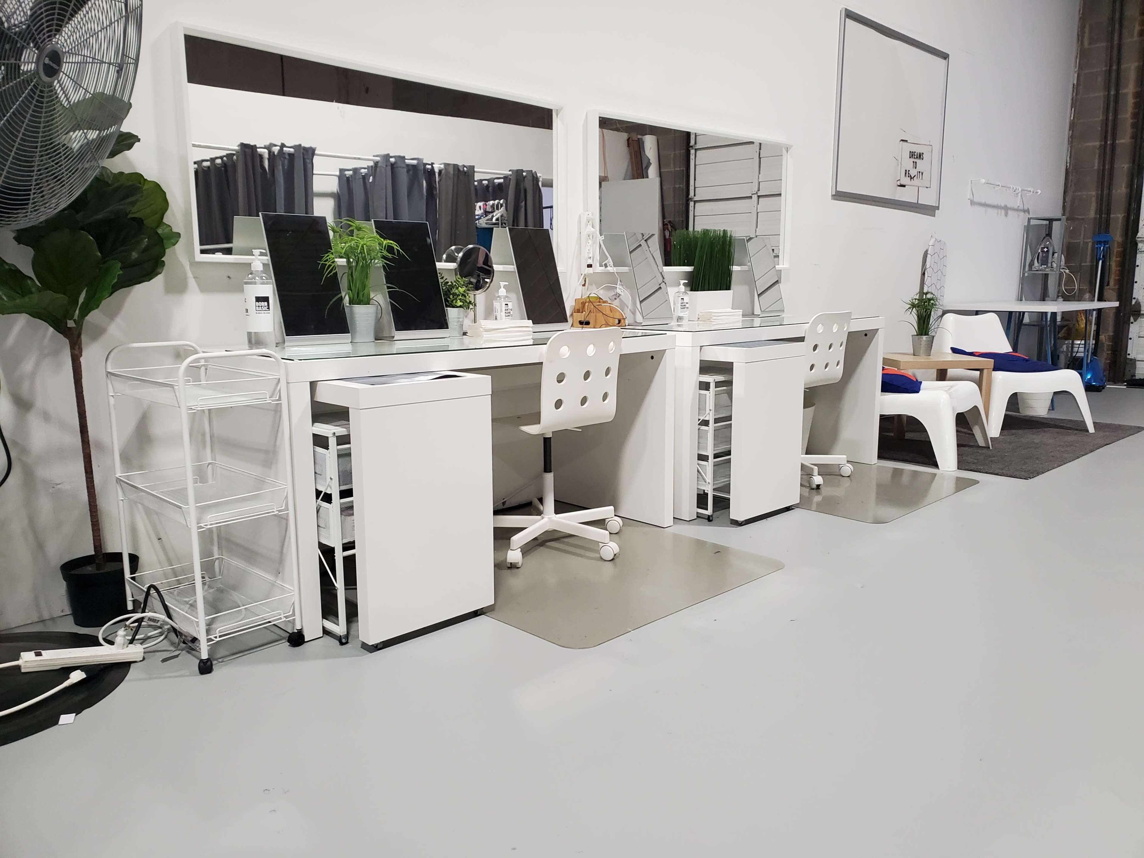 A bright, modern workspace with four identical white desks, each equipped with mirrors and rolling chairs, positioned against a plain wall.