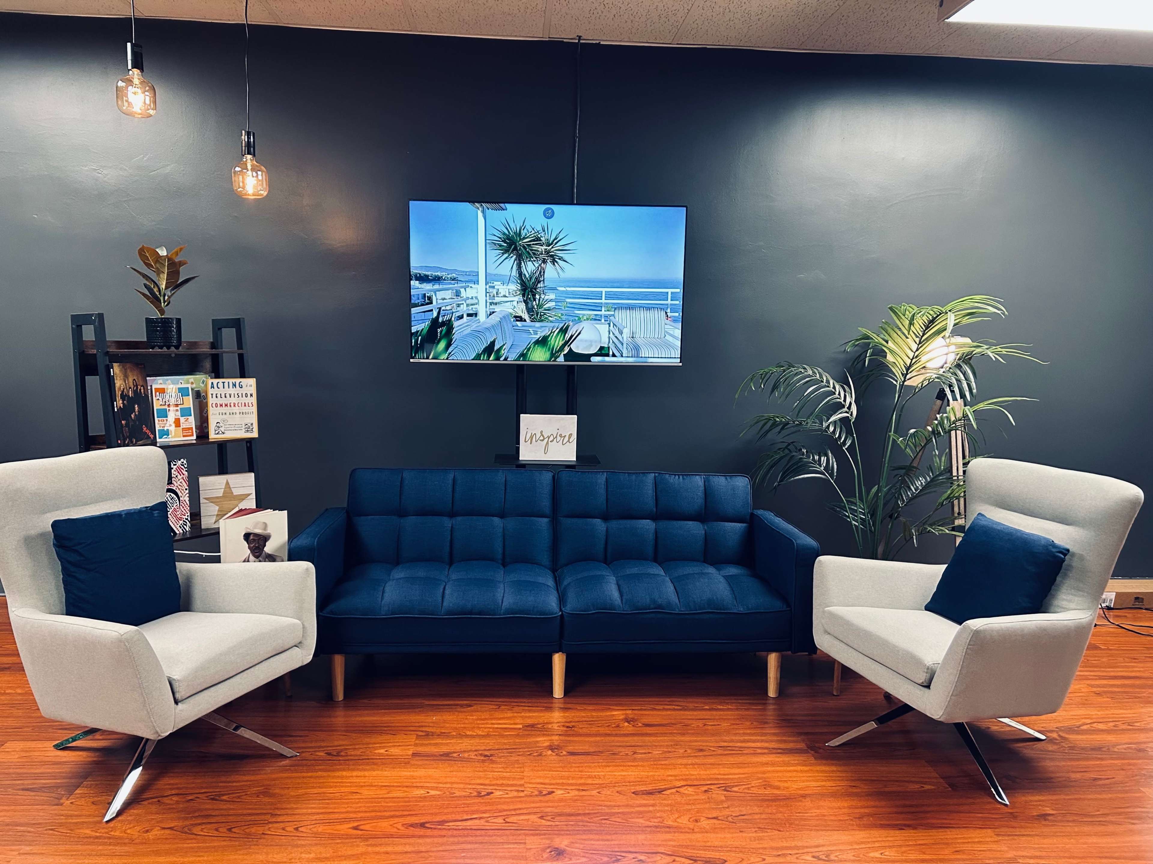 A modern sitting area with a navy blue couch, two light gray armchairs, a television mounted on the wall, and a decorative plant.