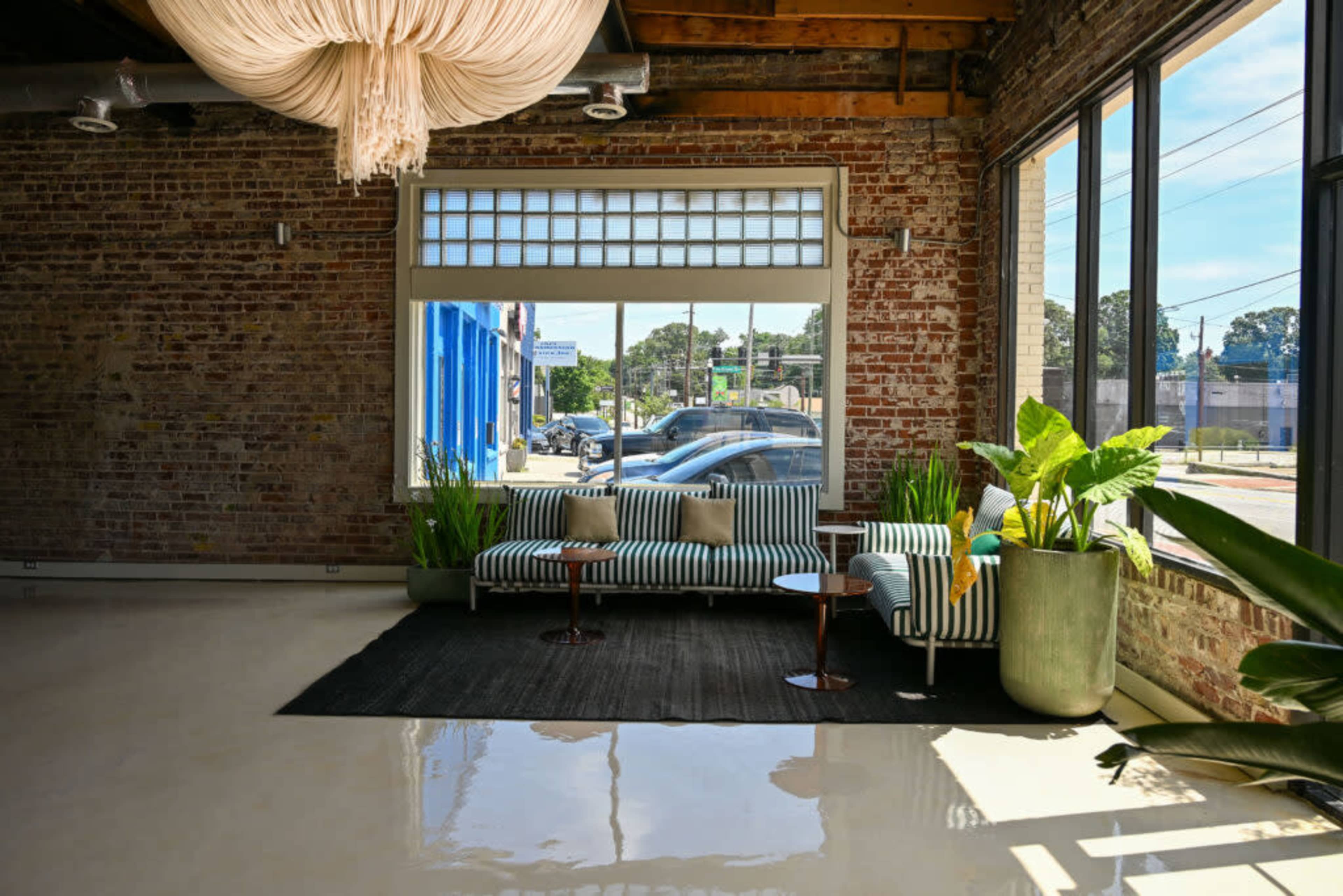 The image shows a modern interior space with a striped sofa and armchair, large windows letting in natural light, and potted plants beside a small table on a glossy floor.