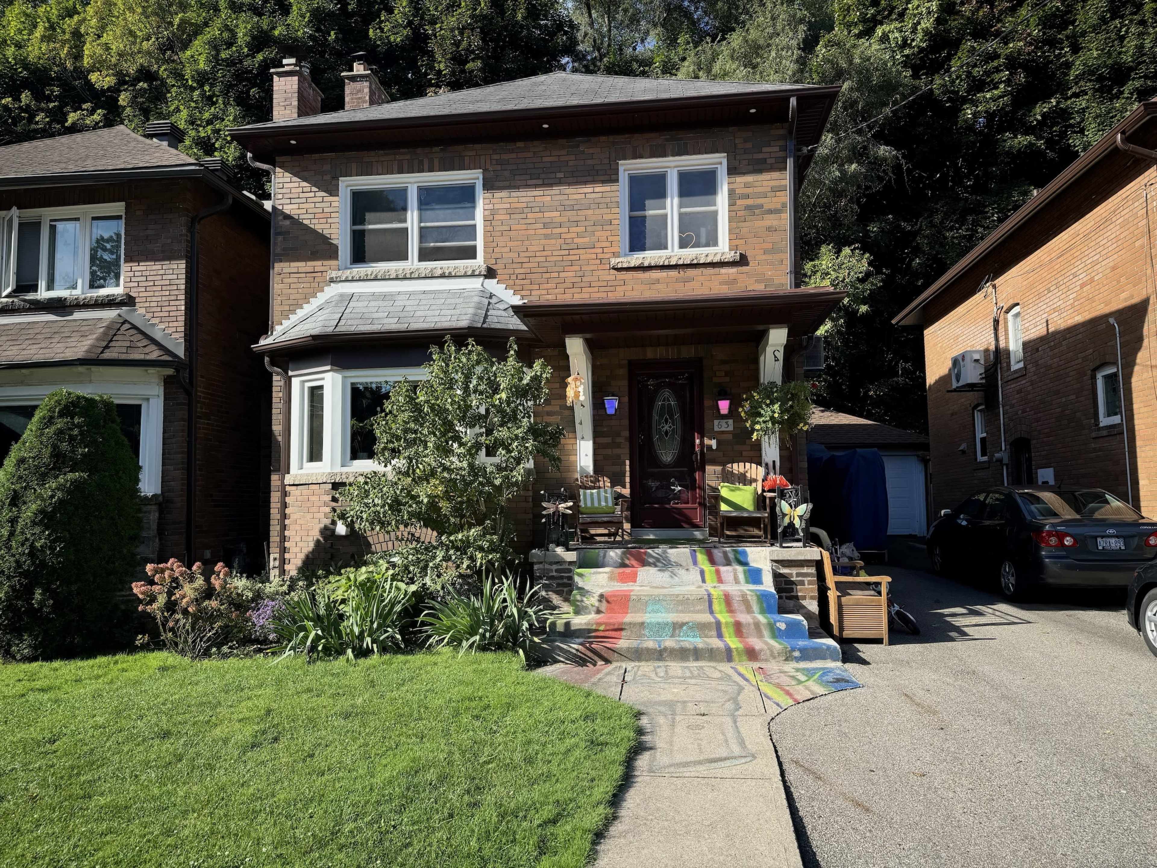 The image shows a two-story brick house with a colorful pathway leading to the front entrance, surrounded by greenery and a parked car nearby.