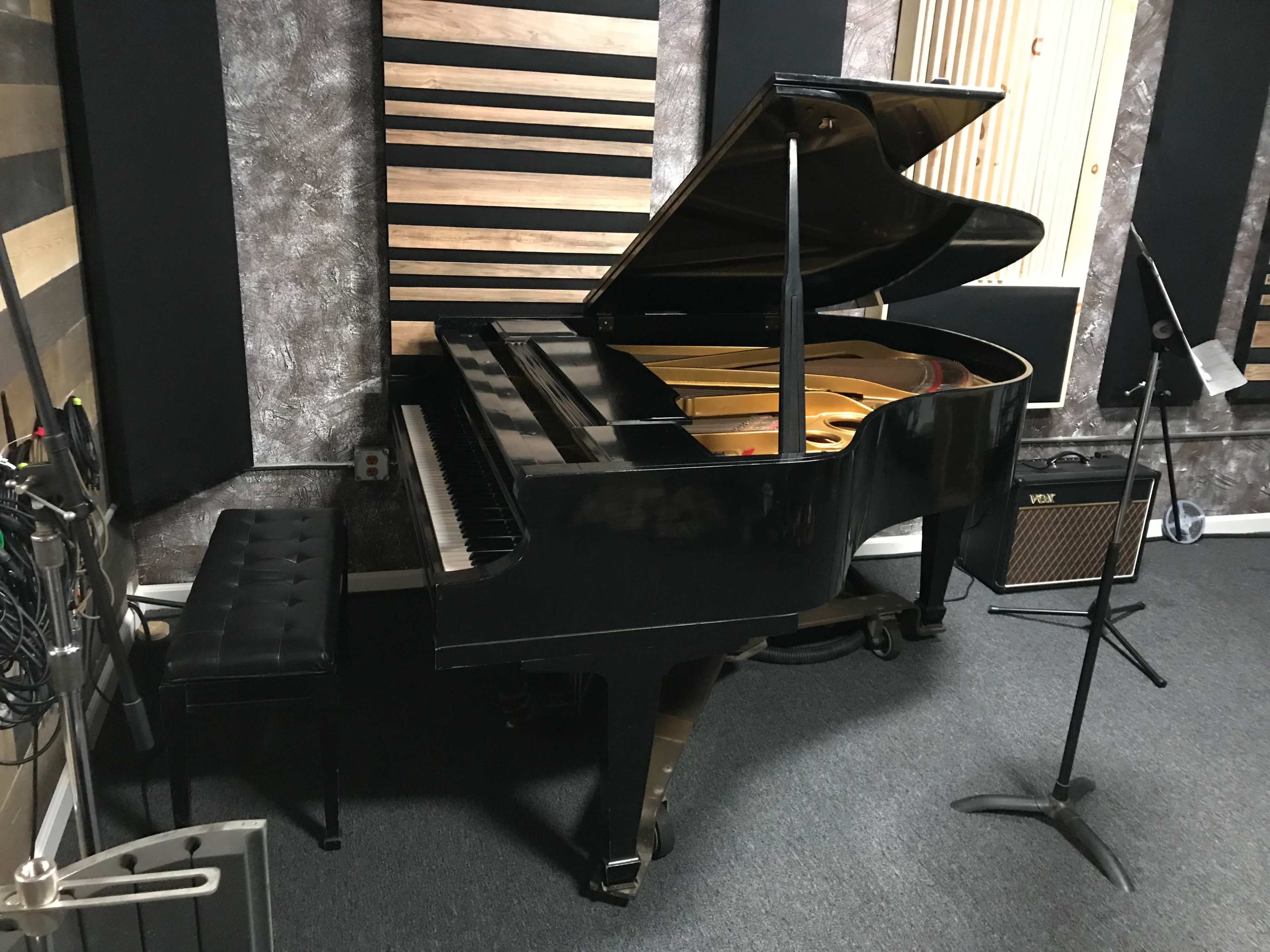 A black grand piano is positioned in a music studio with soundproof panels and a microphone stand nearby.
