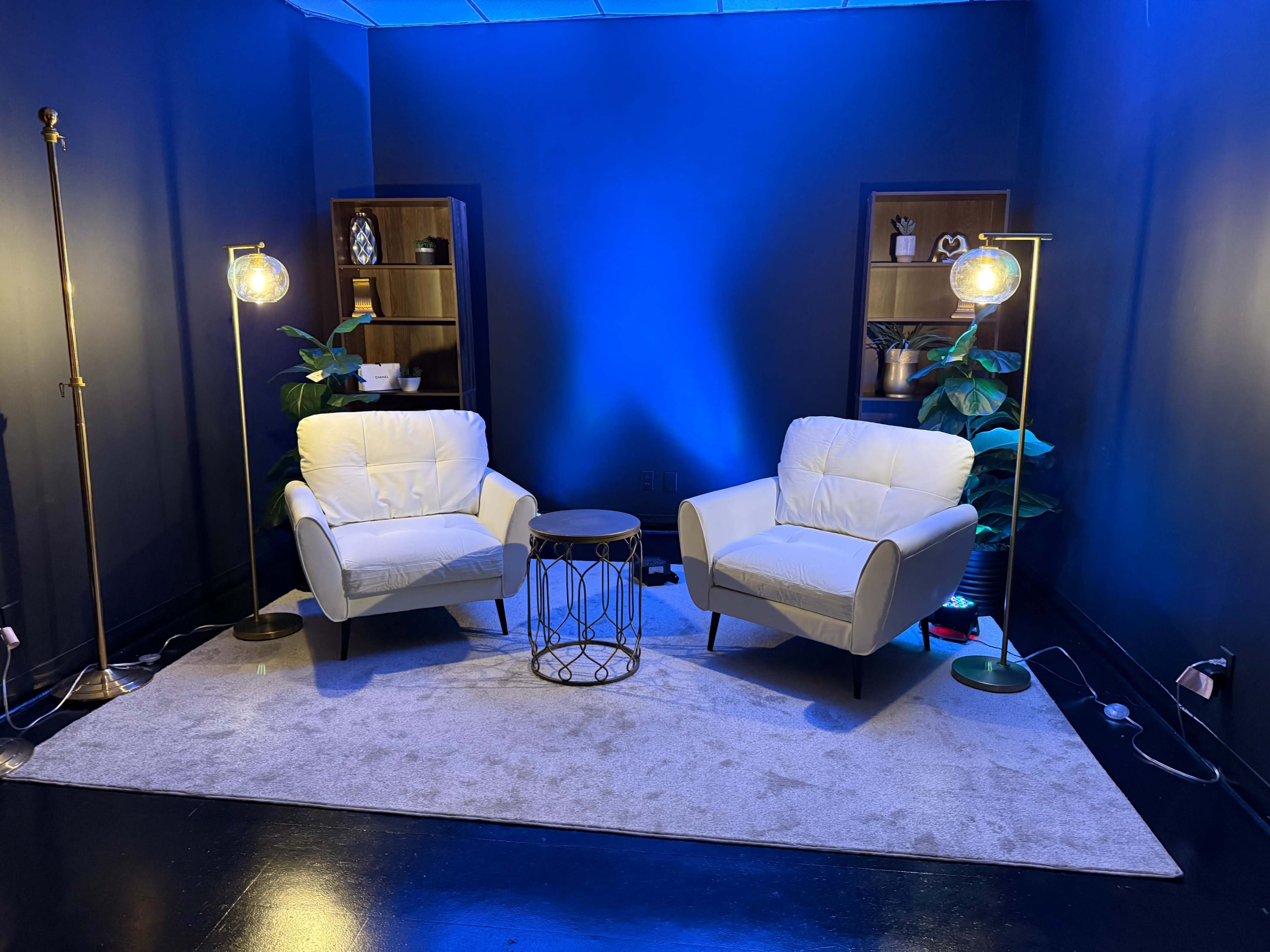 The image shows a cozy seating area with two white armchairs, a small round table, and decorative shelves against a blue-lit wall.