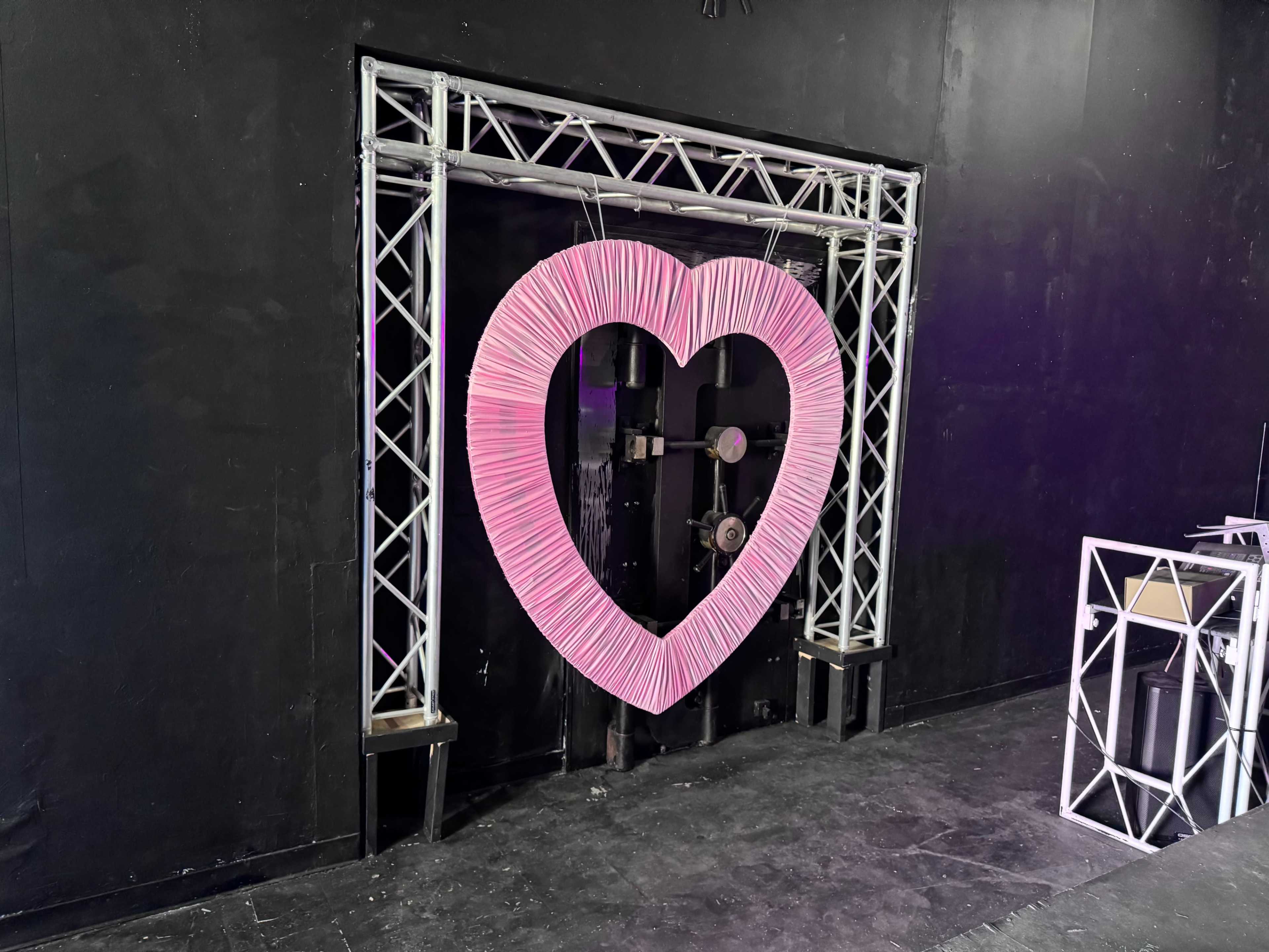 A large pink heart-shaped decoration is suspended in a black walled space, framed by a silver metal structure.