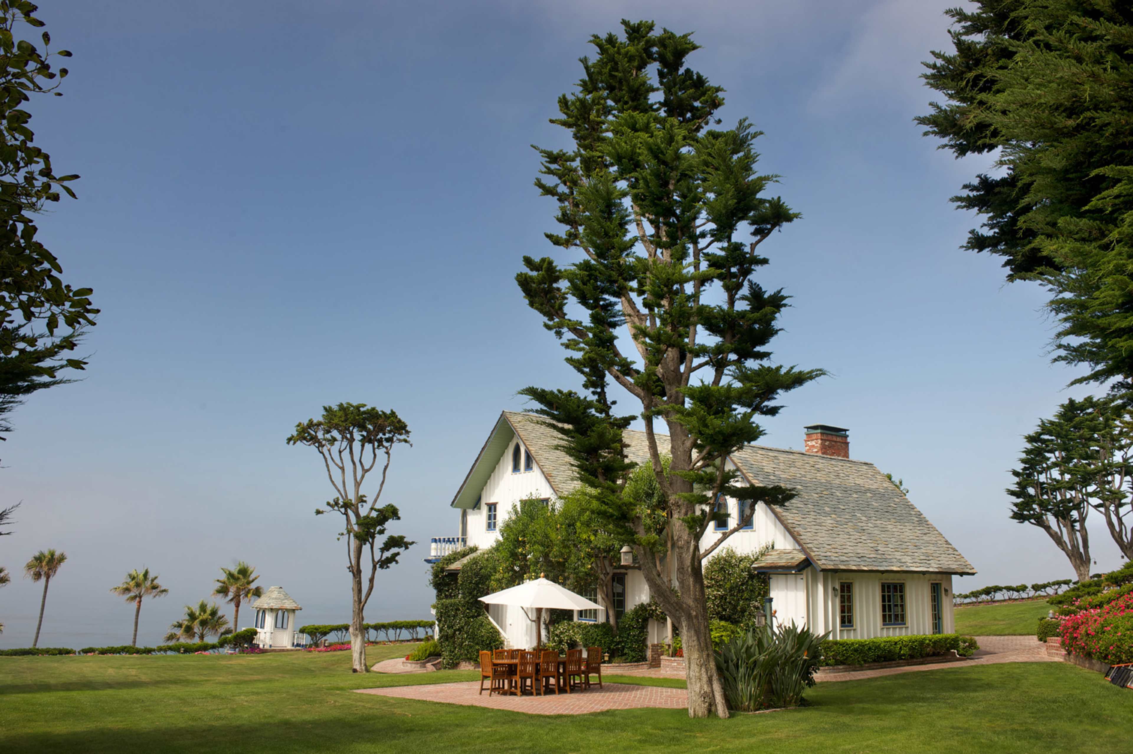A spacious house with a sloped roof is set in a manicured lawn, surrounded by tall trees and palm trees near the coast.