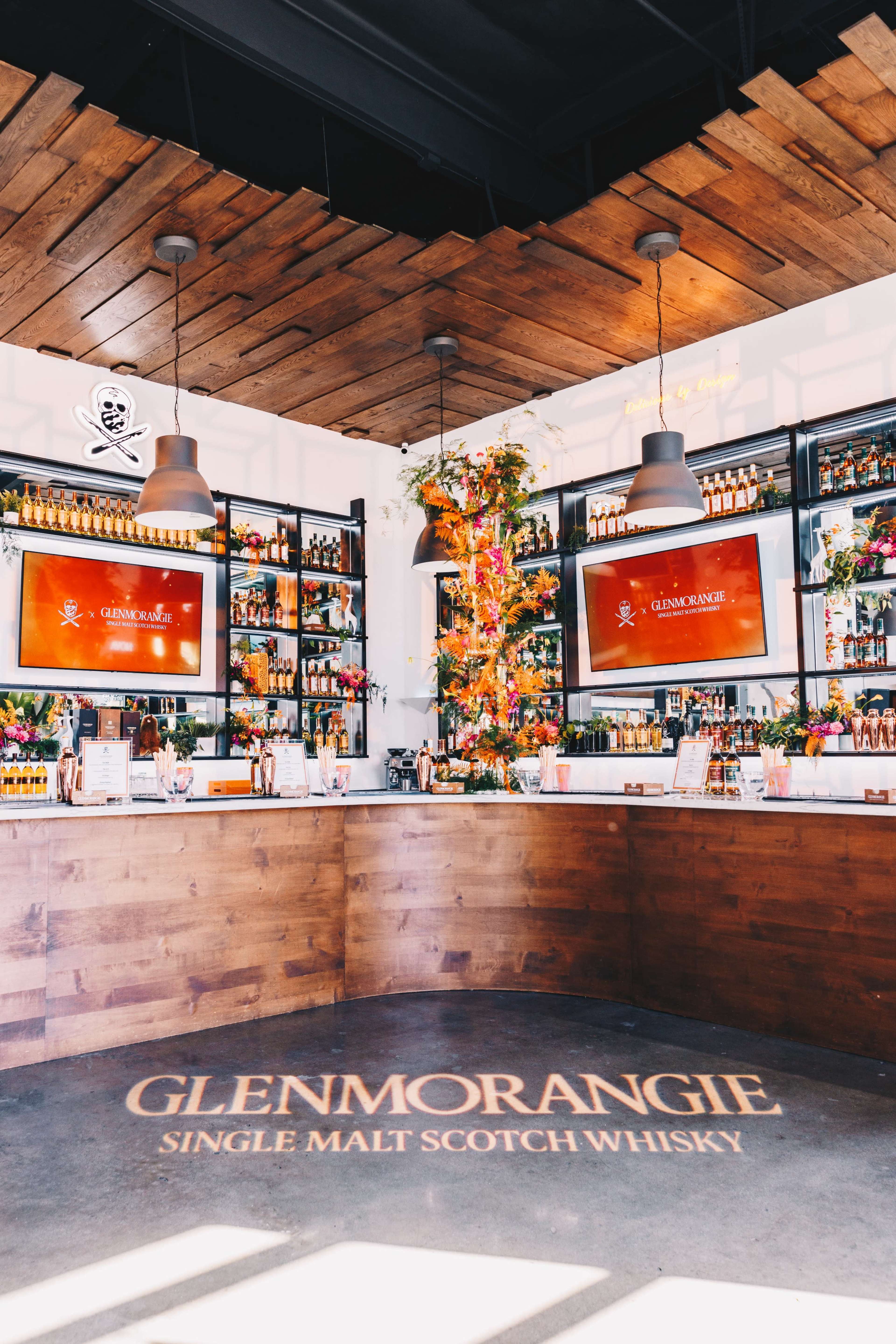 A wooden bar area features displays of Glenmorangie single malt Scotch whisky, with digital screens and colorful floral decorations.