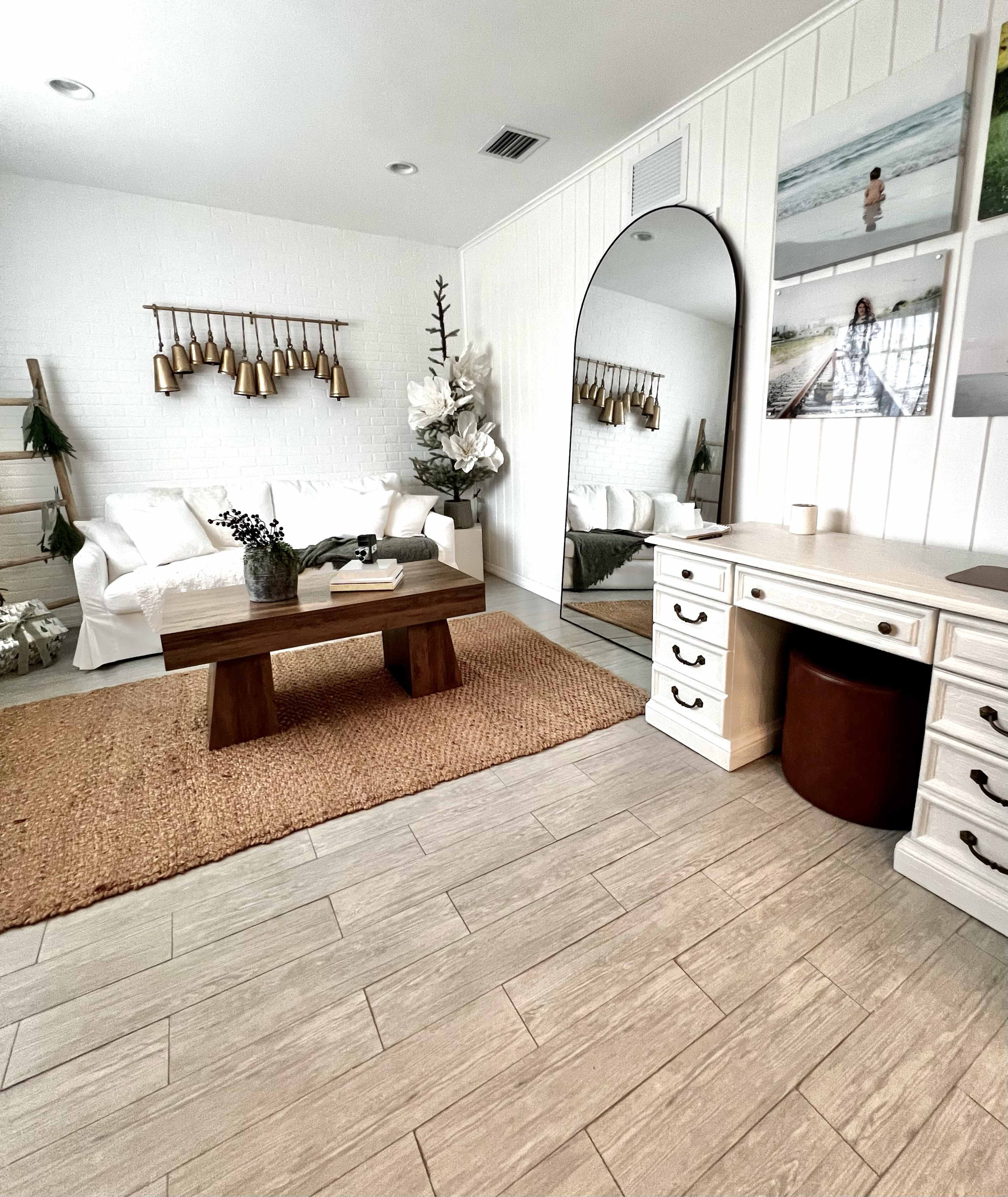 The image shows a modern living room with a white sofa, a wooden coffee table, a large round mirror, and a desk area, all positioned on a light-colored tile floor with a woven rug.