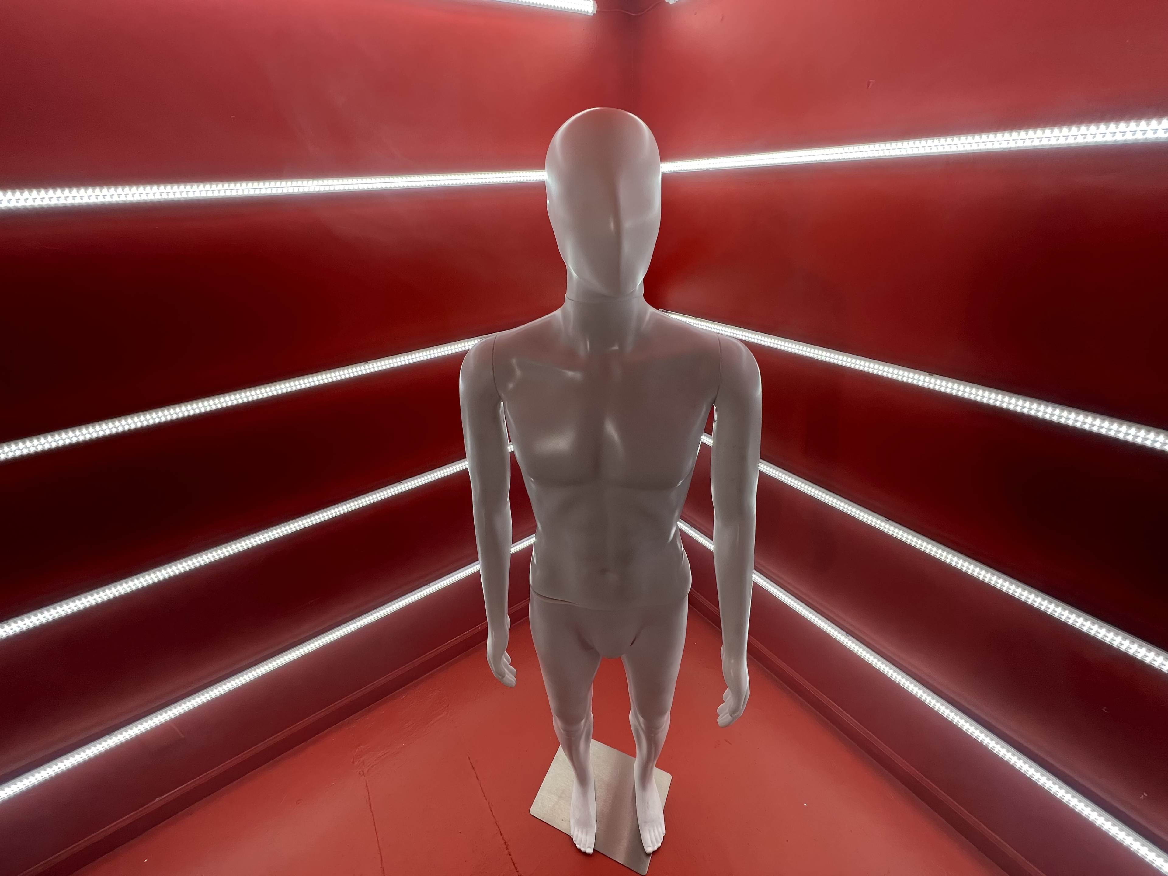 A white mannequin stands in the center of a room with red walls and illuminated by straight lines of LED lights.