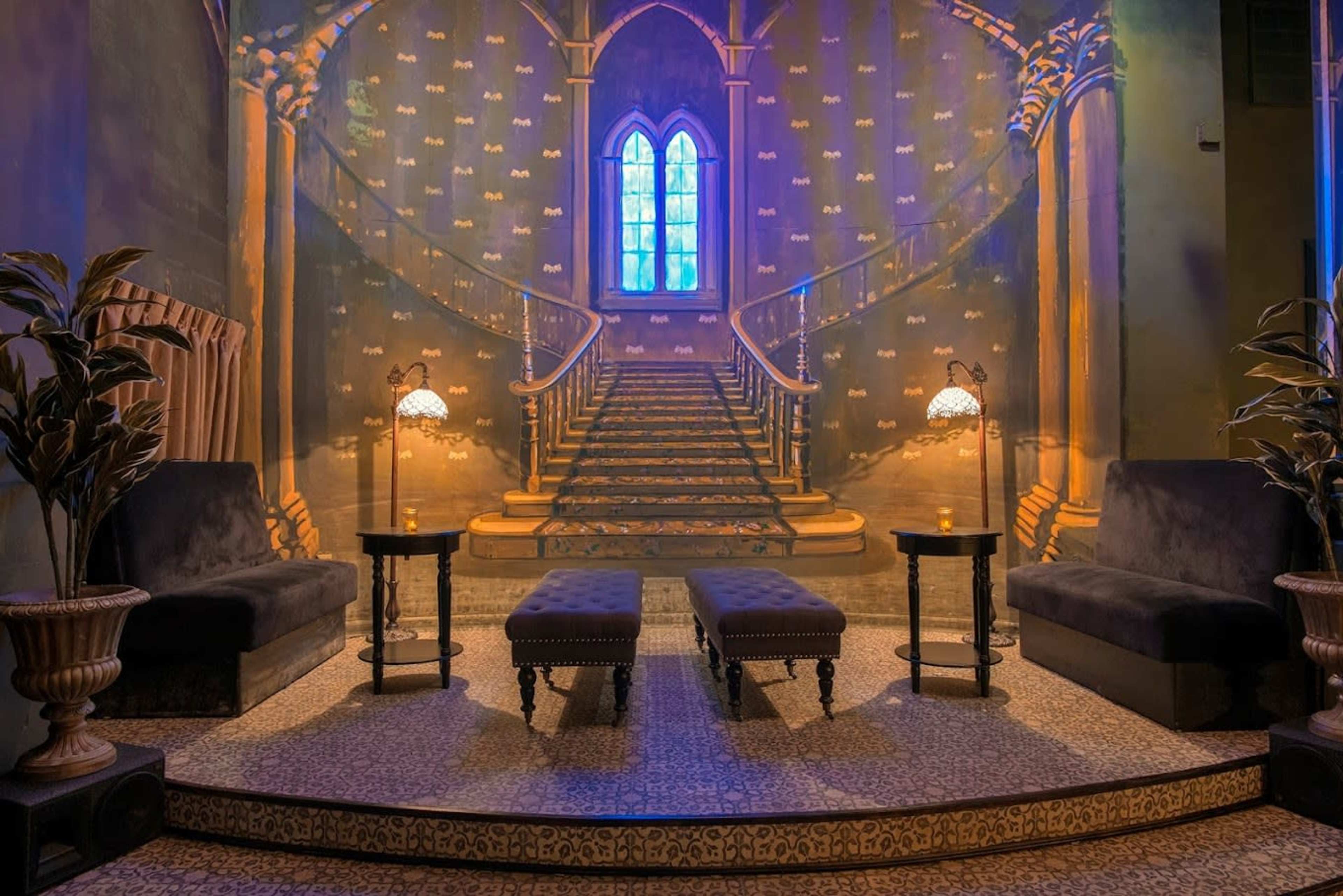 The image features a dimly lit interior with plush seating arranged in front of a large mural depicting a grand staircase and a stained glass window.