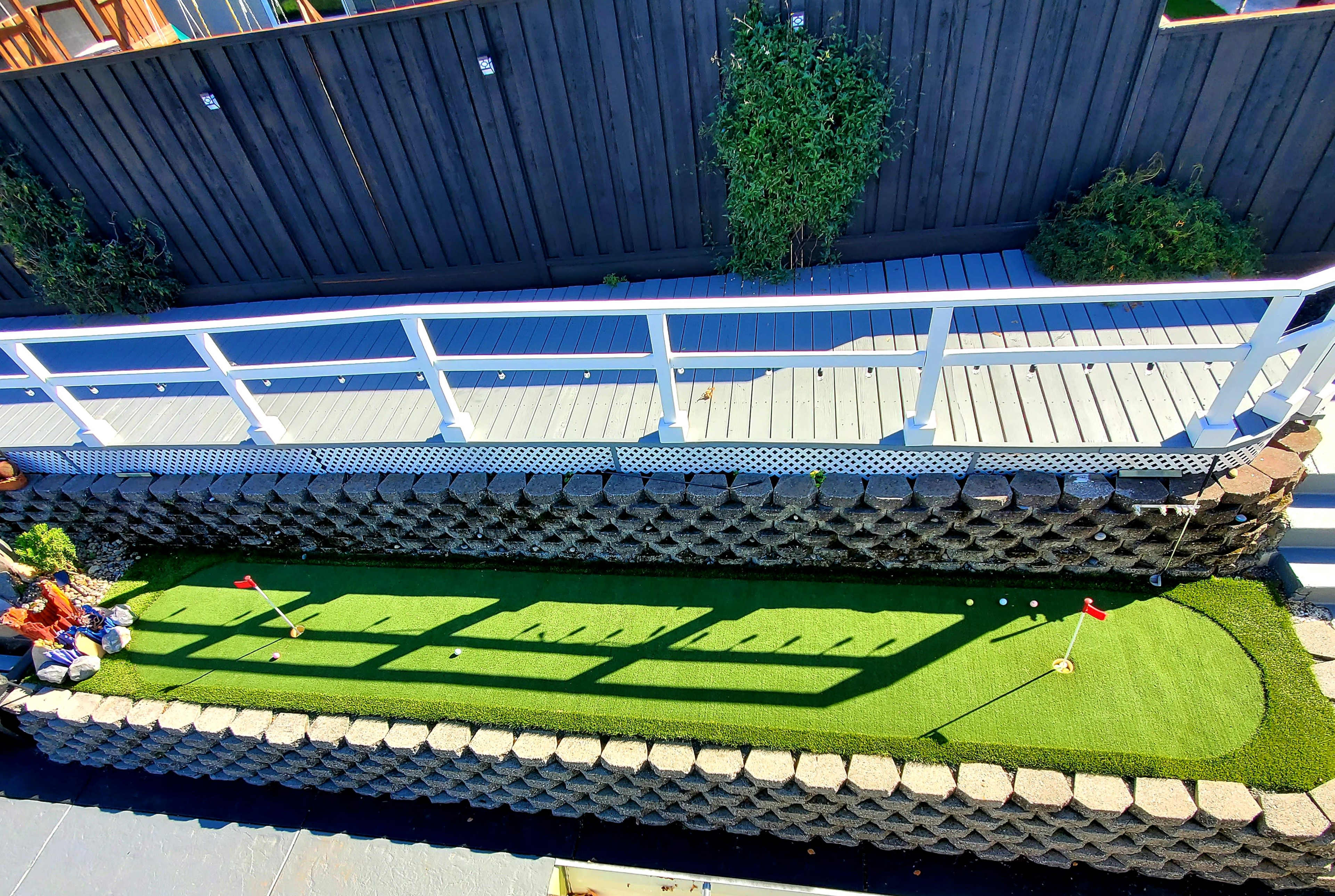 A miniature golf putting green with two flags, surrounded by a stone border, located beneath a wooden deck.