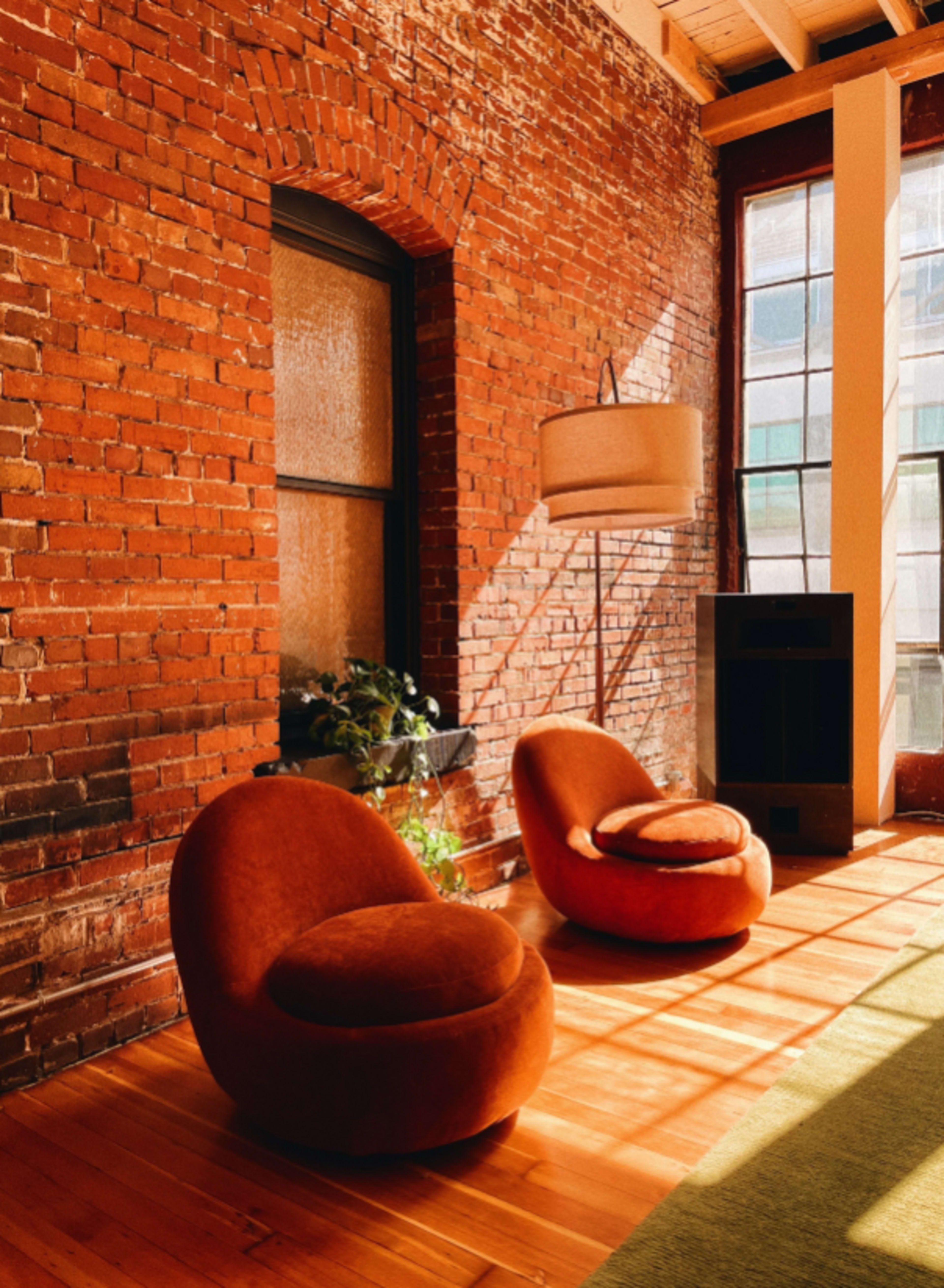 The image shows a cozy corner of a room with two curved orange chairs near a brick wall, complemented by a standing lamp and large windows allowing natural light to flood the space.