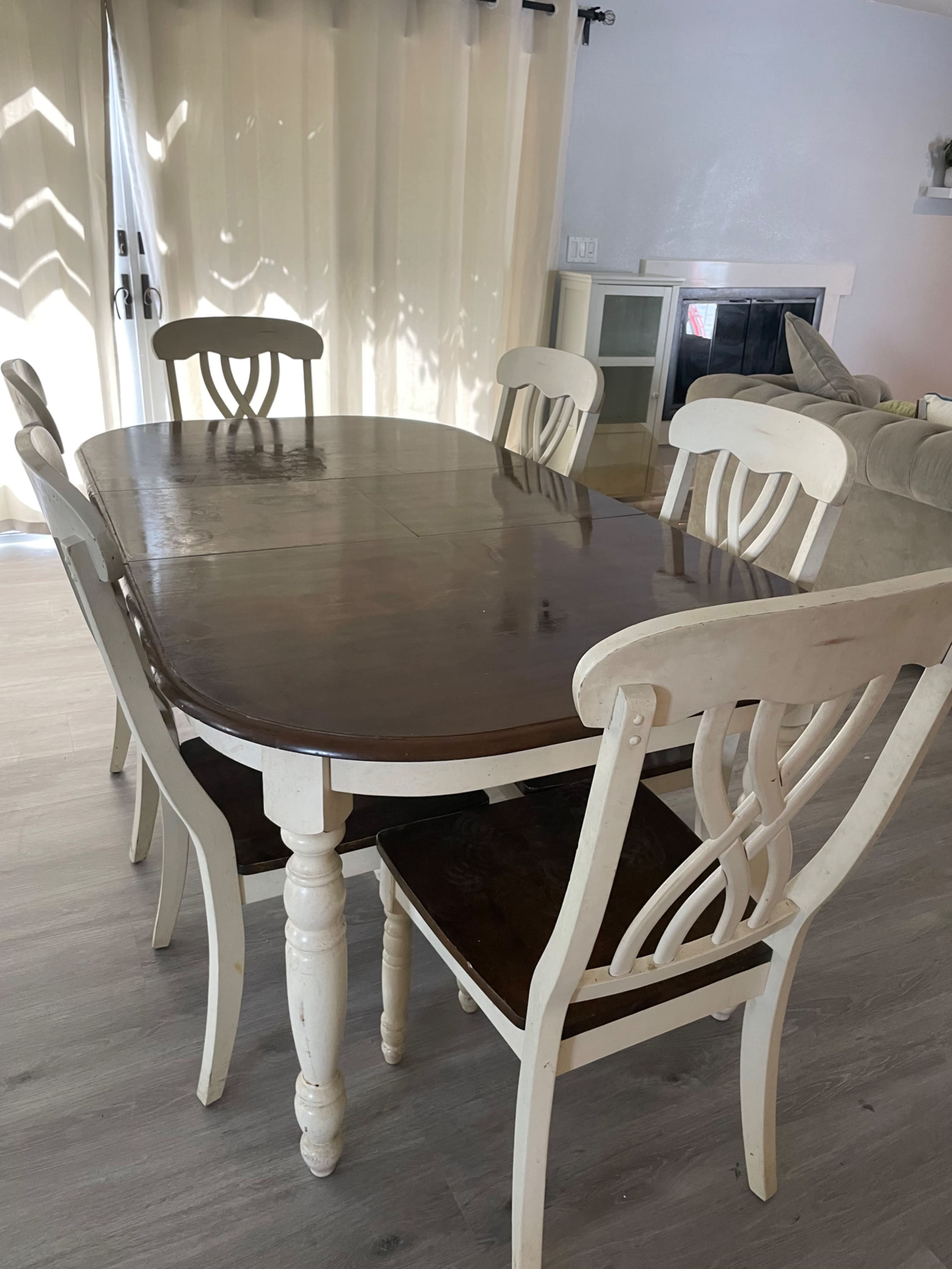 A wooden dining table with six chairs is set in a well-lit room with light-colored walls and large windows.