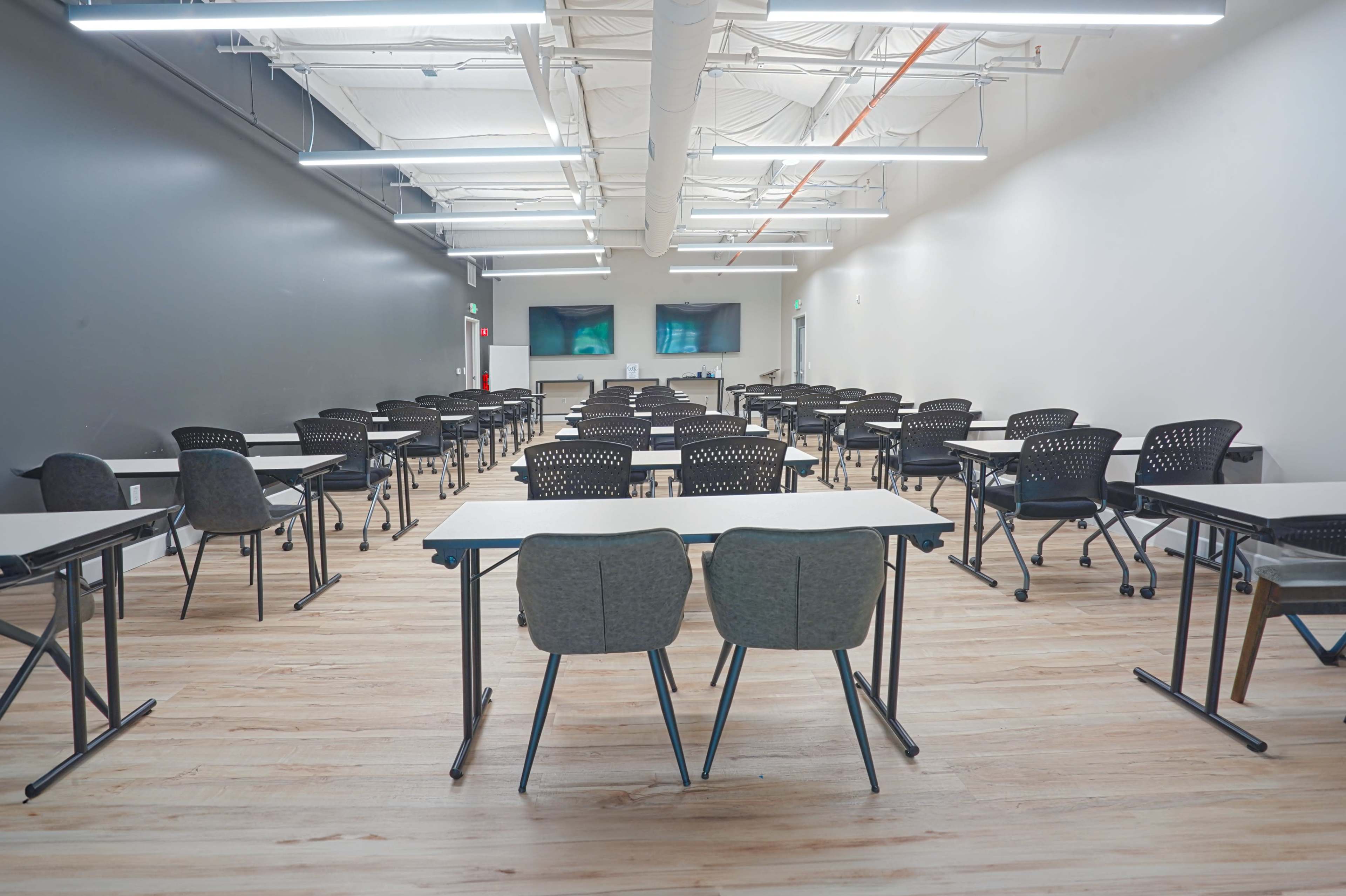 A classroom features rows of desks and chairs arranged neatly on a wooden floor, with two screens mounted on the wall at the front.