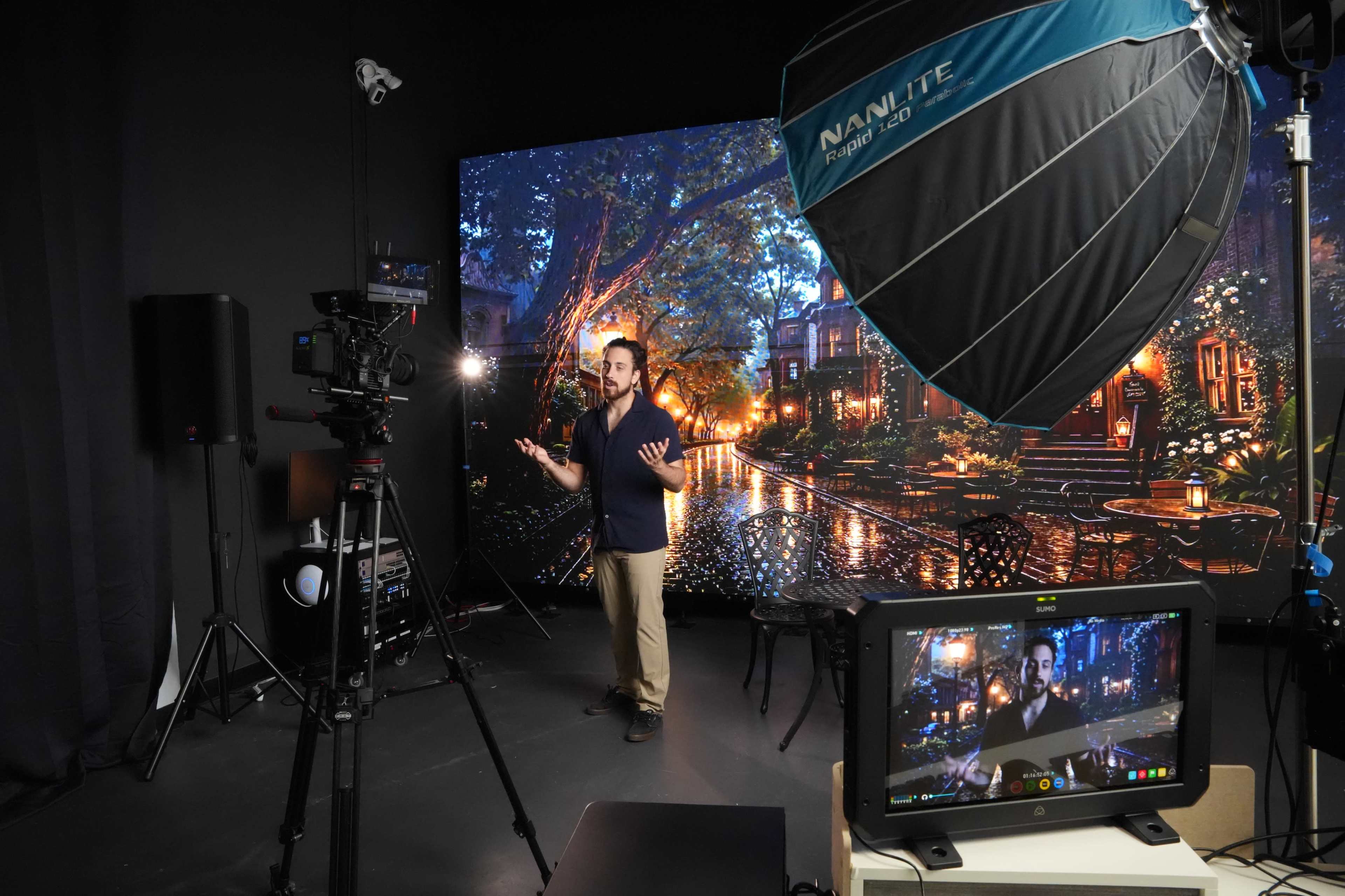 A man stands in front of a large backdrop depicting a nighttime outdoor scene, with studio equipment visible around him.