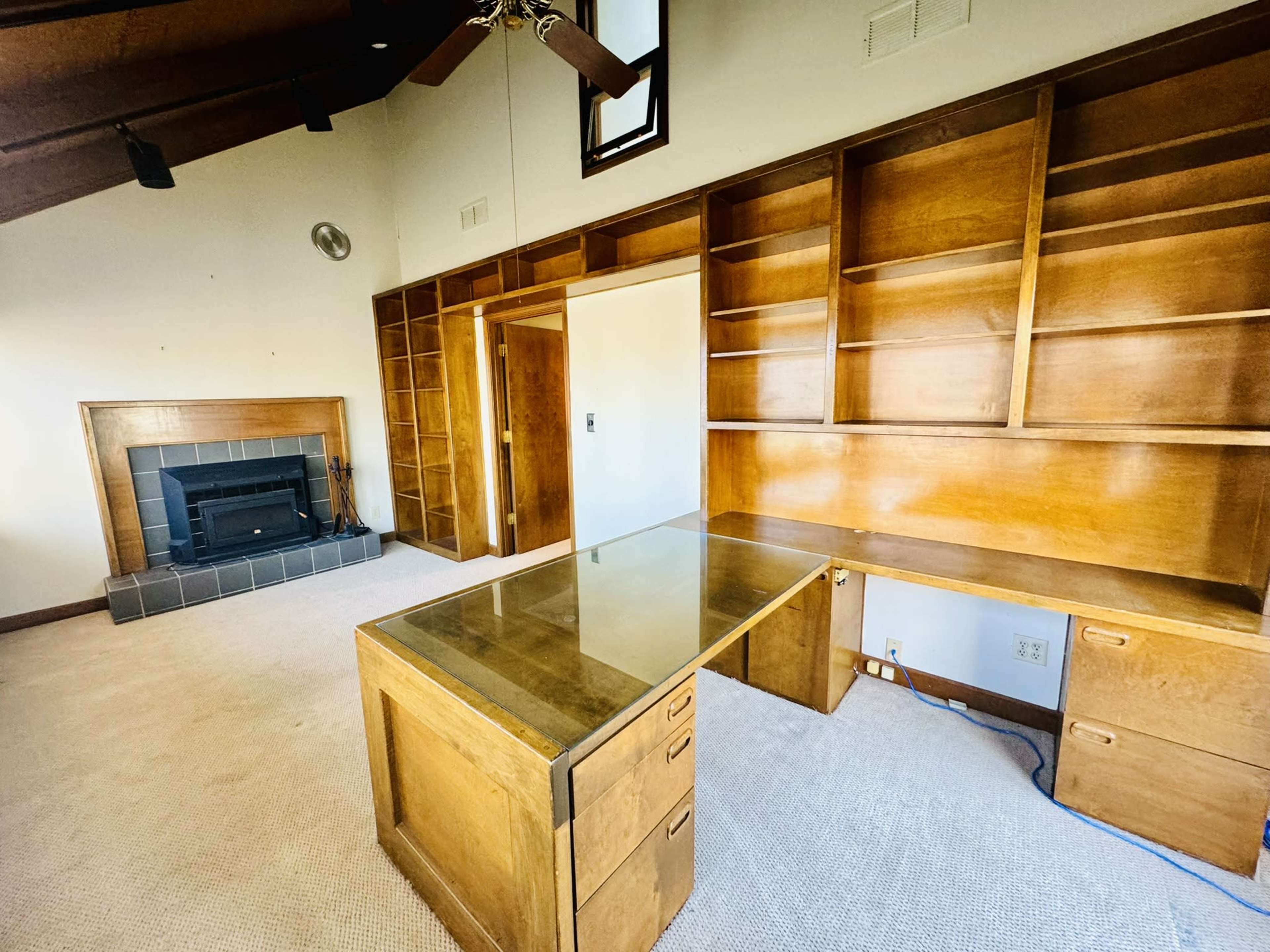 A room with a wooden desk and shelving unit against a wall, featuring a fireplace and a high ceiling.
