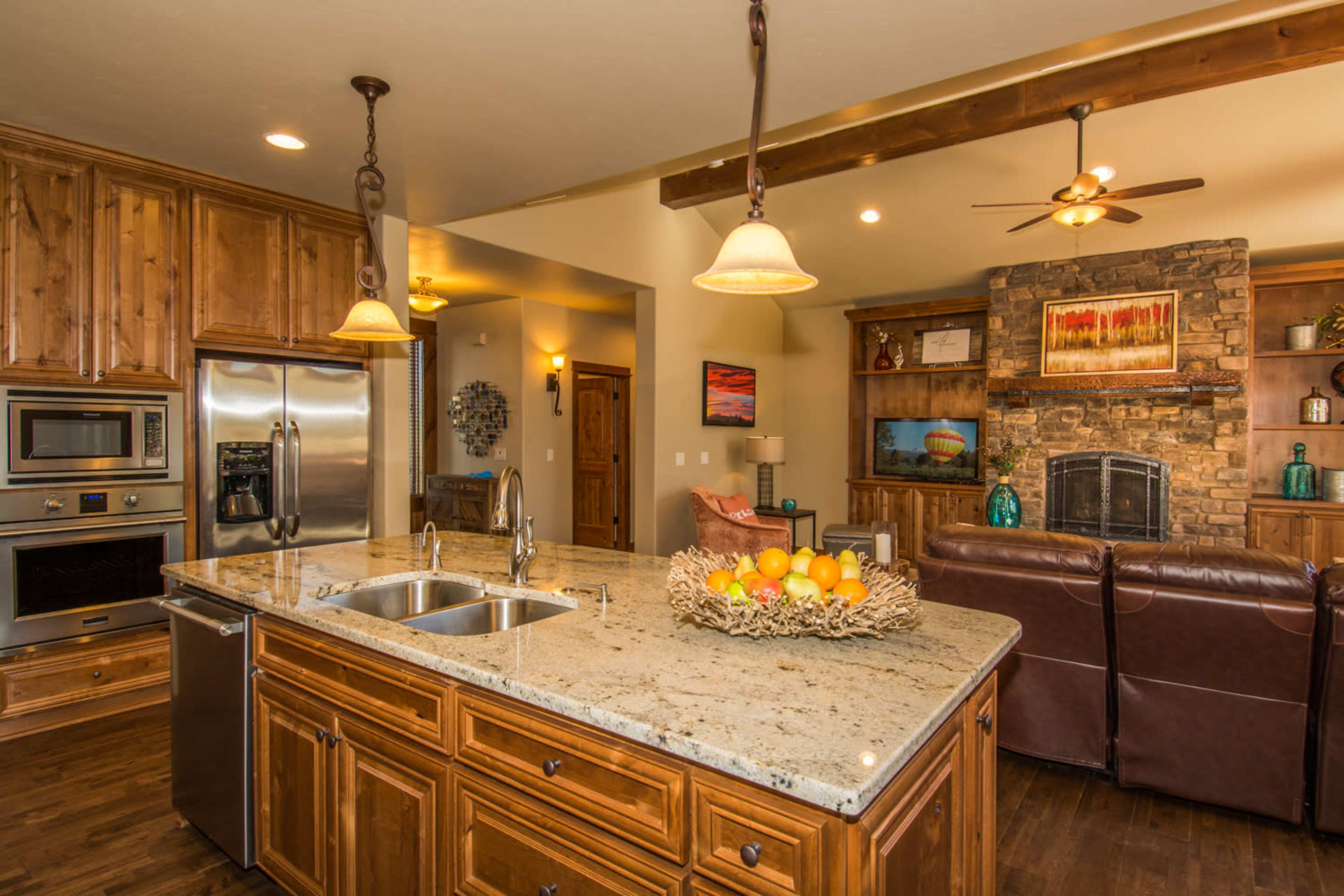 The kitchen features wooden cabinetry, a central island with a double sink, and a living area with a stone fireplace in the background.