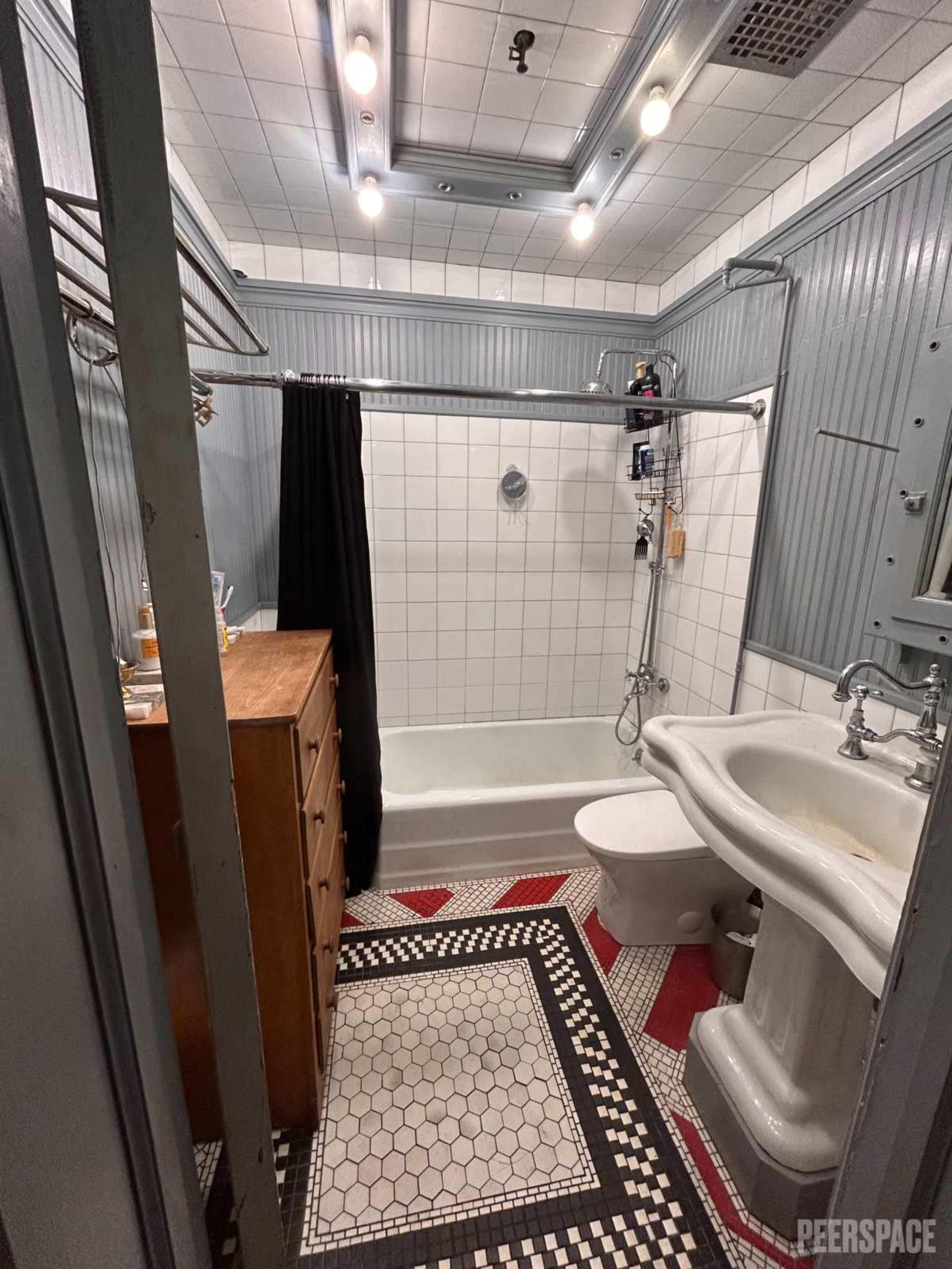 The bathroom features a bathtub with a shower, a sink, and a patterned tile floor, along with a wooden dresser and various bathroom fixtures.