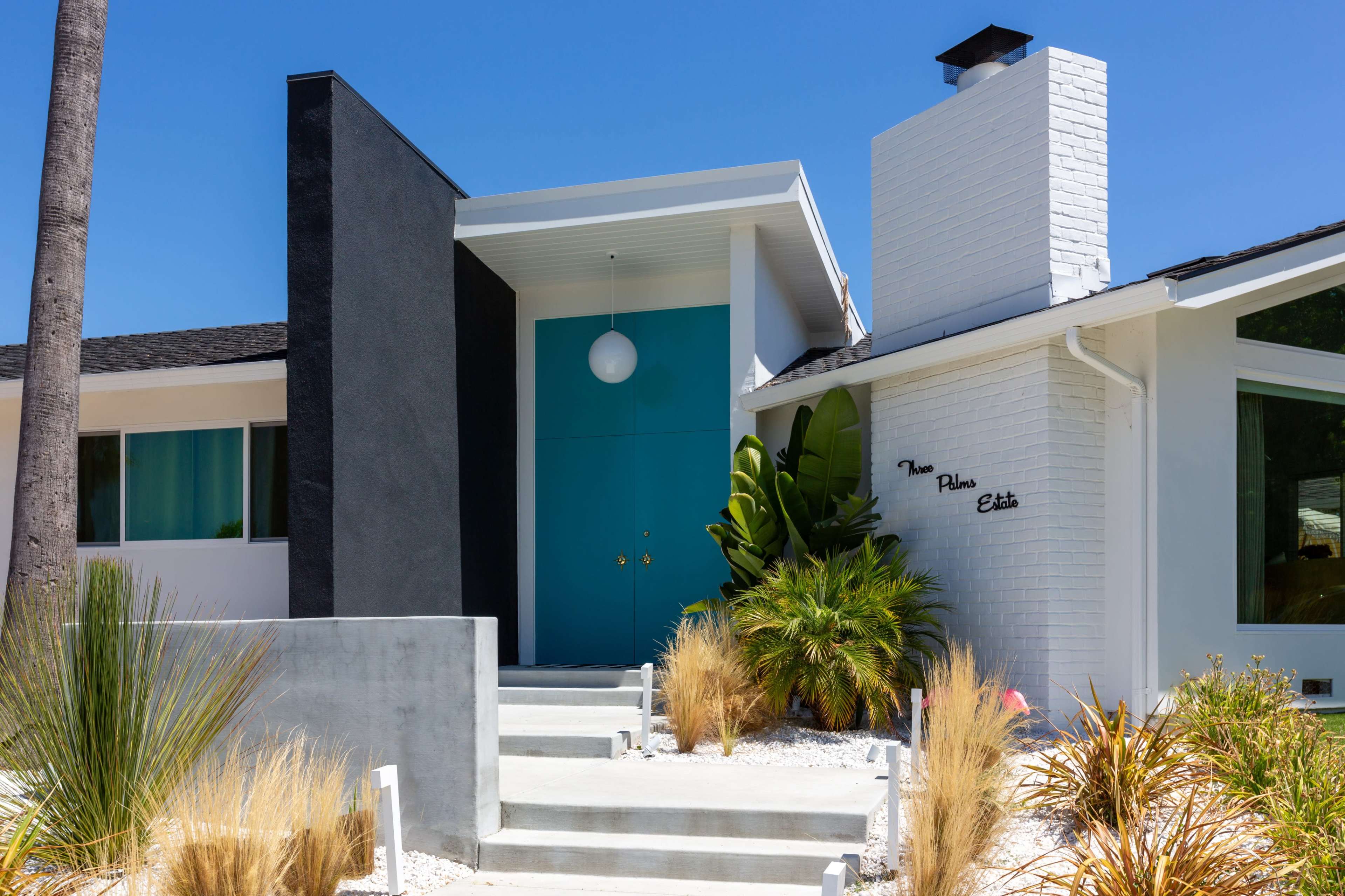 A modern house features a contrasting black wall, a bright turquoise door, and a landscaped front yard with various plants.