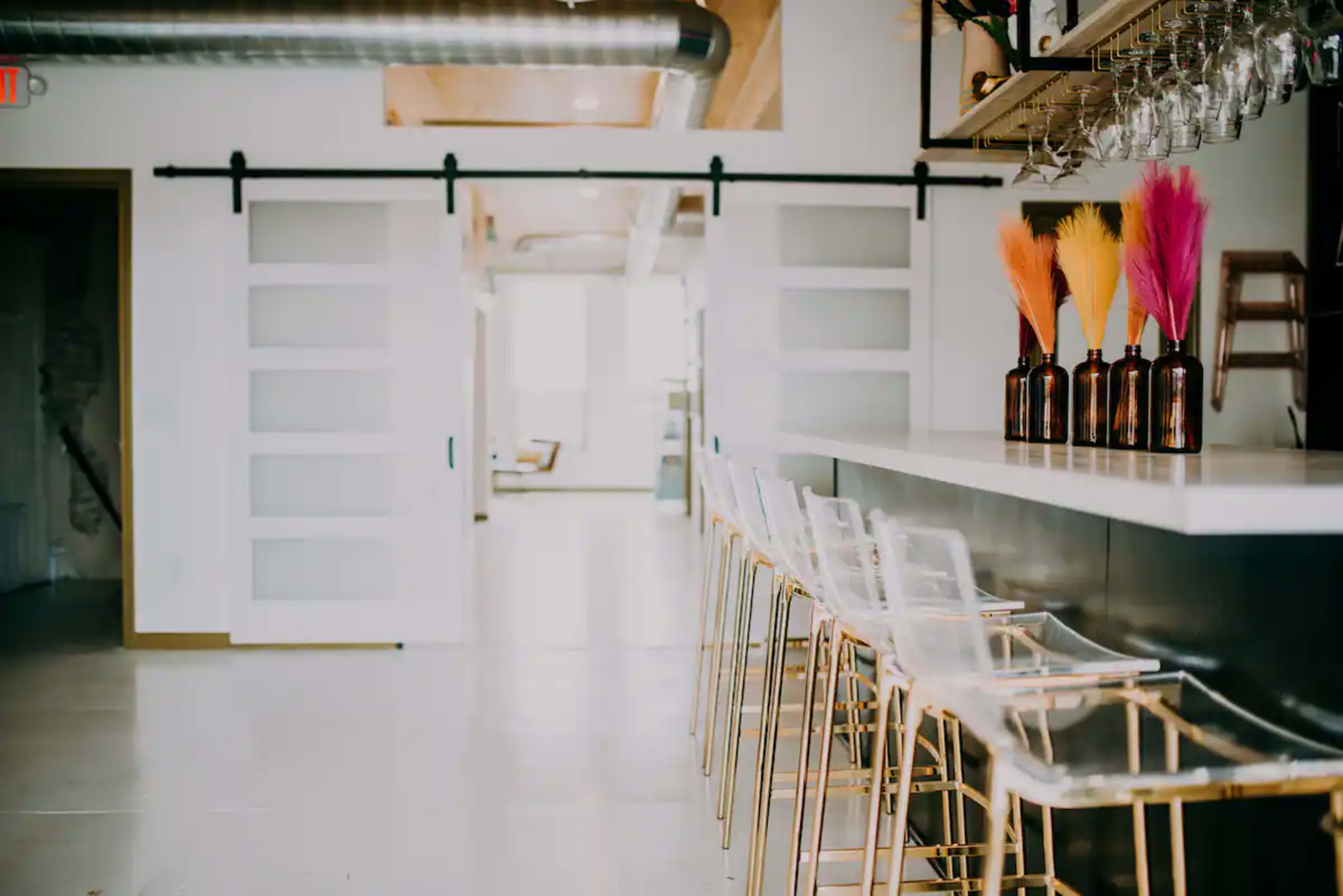 A modern bar area with clear stools and colorful decorative vases on the counter, flanked by sliding doors leading to a bright space.
