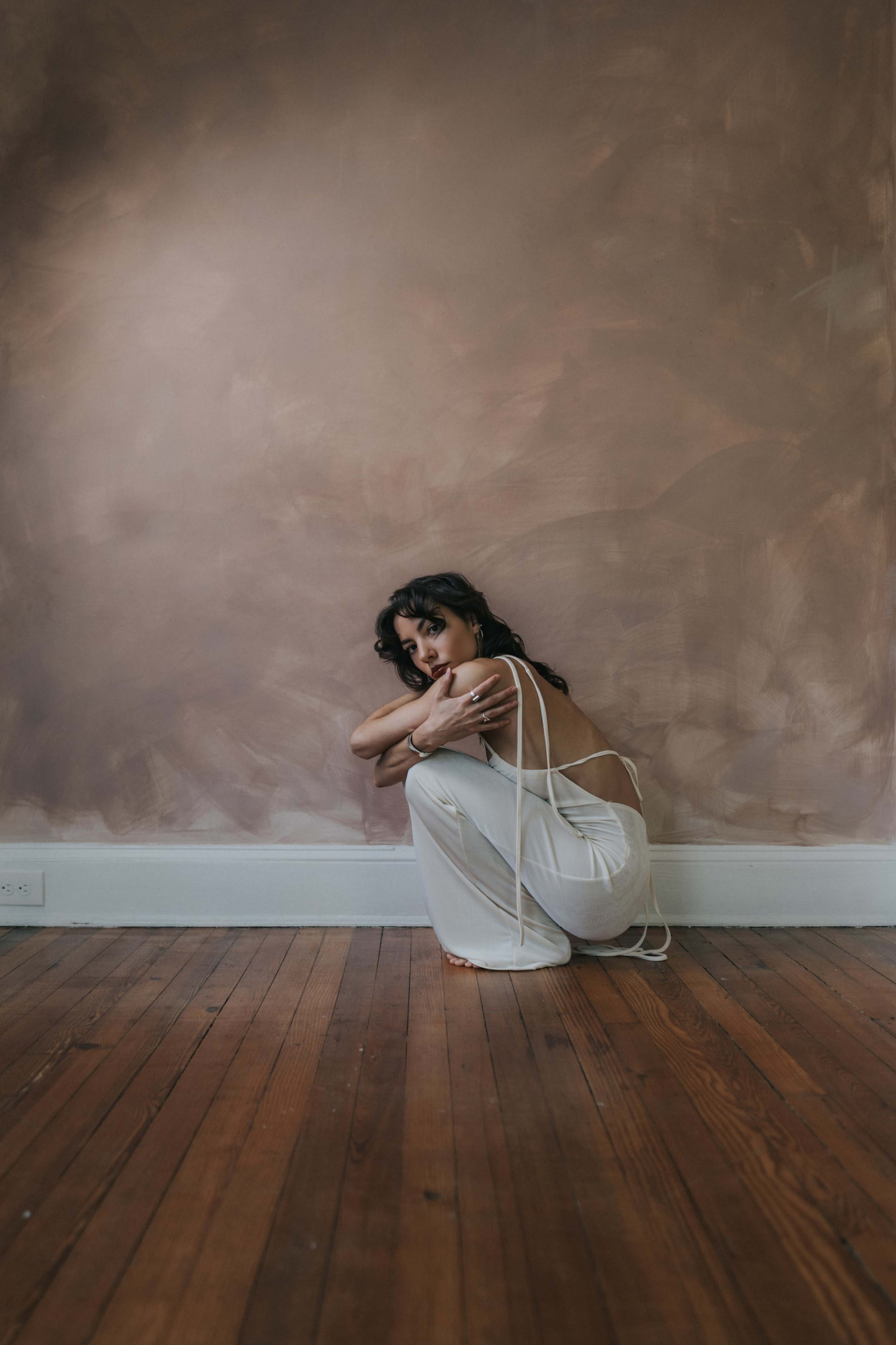 A woman in a white outfit is sitting on a wooden floor with her arms wrapped around her knees against a softly painted wall.